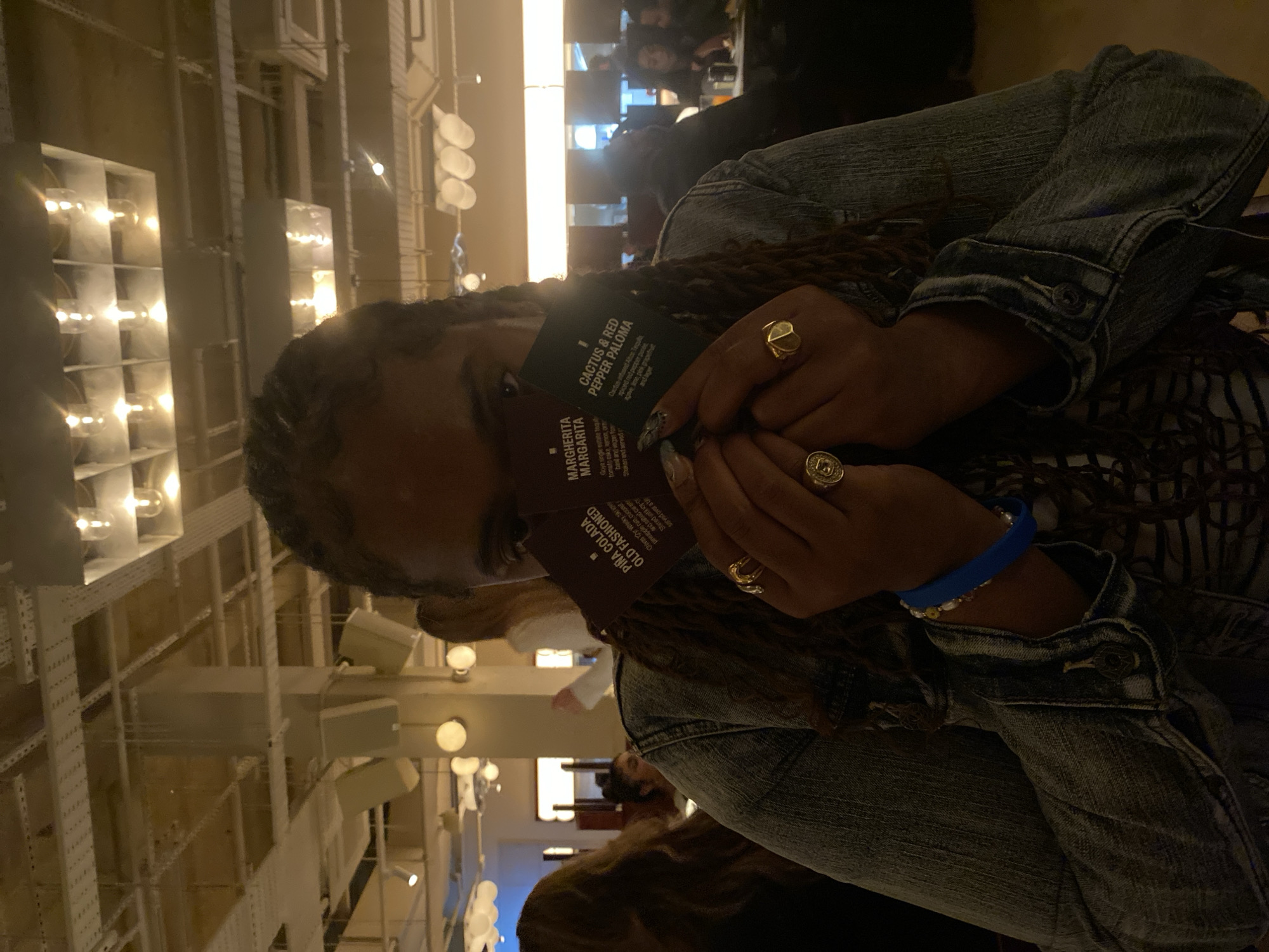 A woman with long, braided hair is in a dimly lit restaurant, holding up three drink menus: "Margherita Margarita", "Piña Colada Old Fashioned", and "Cactus & Red Pepper Paloma". She wears a denim jacket, and has multiple rings on her fingers and a blue wristband. The restaurant interior has a modern industrial design, with visible pipes and lighting fixtures. Other people can be seen in the background, implying a social setting. The location cannot be identified based on the visual information provided.