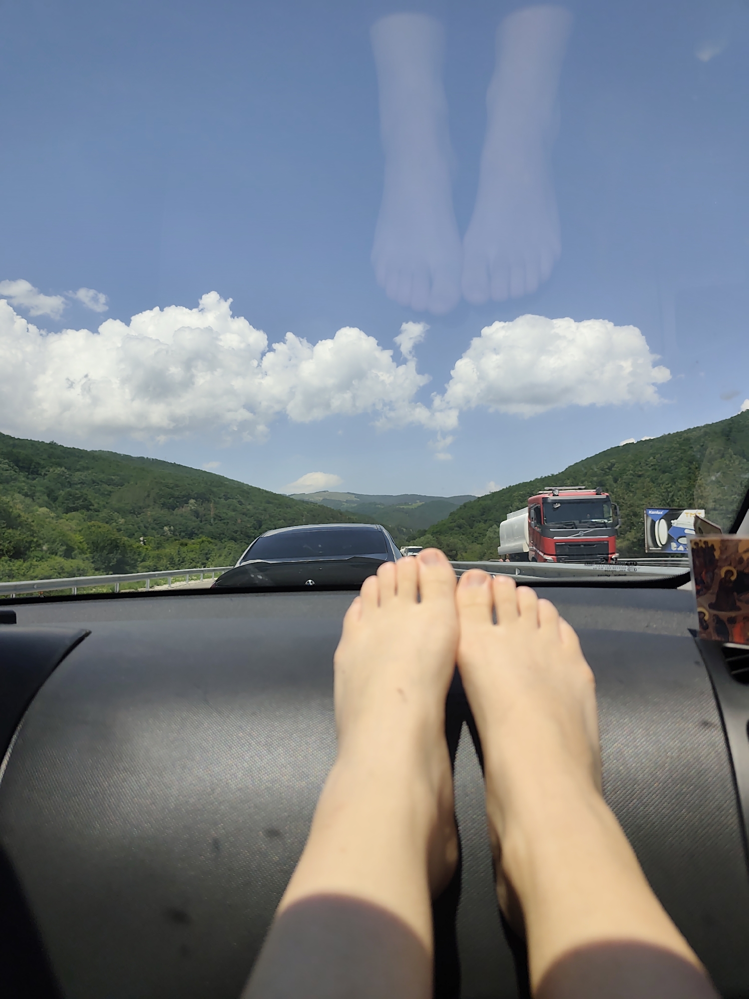 The image captures a first-person perspective from the interior of a vehicle, looking through the front windshield on a clear, sunny day. In the immediate foreground, two bare feet with light skin are positioned side-by-side on a dark, textured dashboard. Above the feet, through the windshield, a multi-lane highway is visible, receding into the distance. A dark-colored vehicle travels directly ahead, partially obscured by the dashboard. Further down the road on the right, a red and white tanker truck is visible, accompanied by a roadside billboard featuring "Kanlux" and "TMT" branding. Lush green, tree-covered hills and mountains dominate the midground and background, extending towards a distant horizon. The sky is bright blue with numerous white cumulus clouds. A faint, ghostly reflection of the two feet is distinctly visible in the upper portion of the windshield, superimposed against the sky. A small, dark object with a reddish-brown, possibly ornate, pattern is partially visible on the far right of the dashboard. The location is an unidentifiable rural or semi-rural highway, not within a city.
