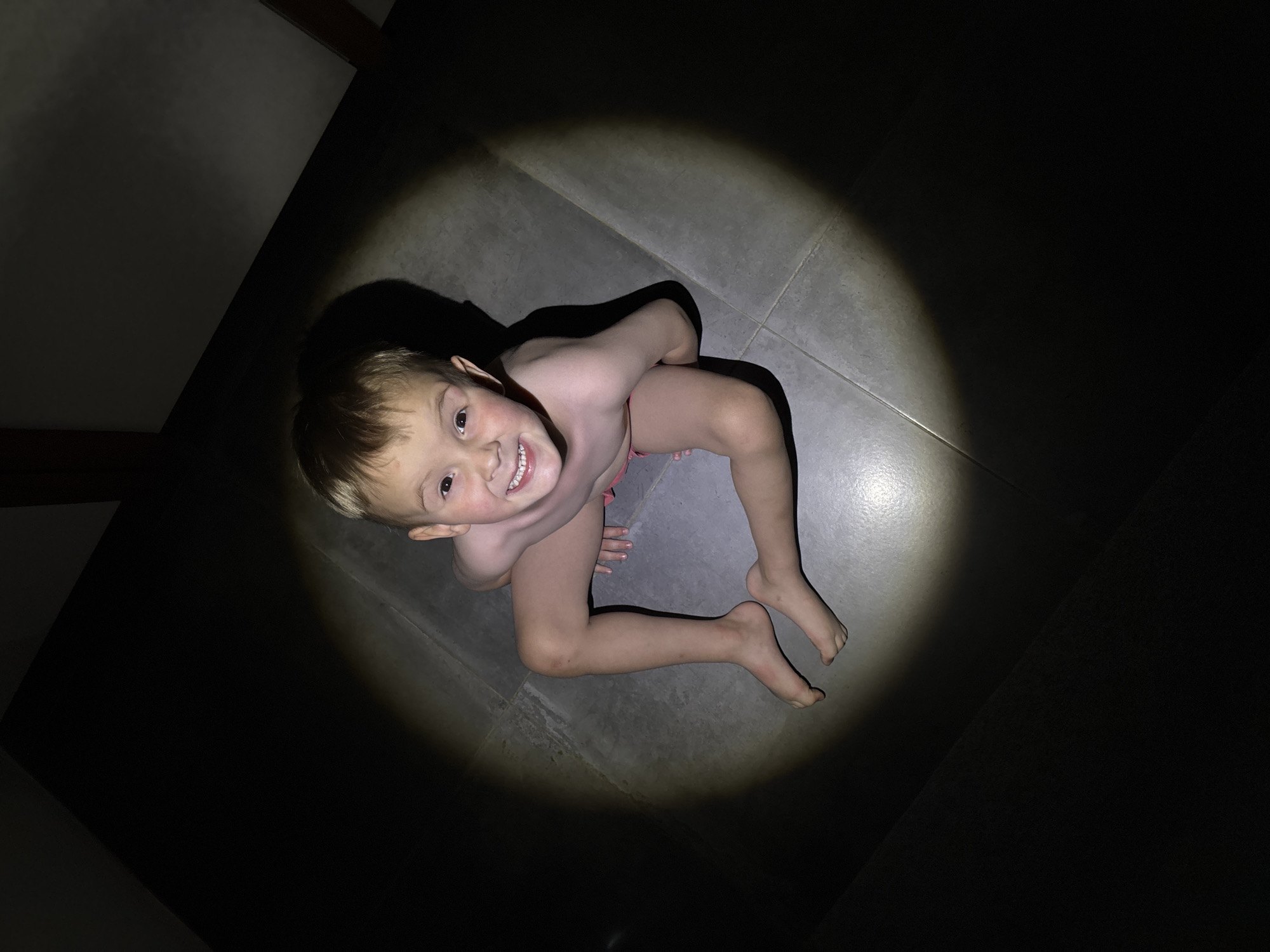 A high-angle shot depicts a young, fair-skinned child illuminated by a circular light source against a dark background, which appears to be a room in a building. The child, positioned on a grey-tiled floor, is smiling and looking up. They appear to be wearing minimal clothing, possibly just underwear, and their skin shows light and shadows from the lighting. The surrounding area is mostly dark, with only portions of the floor illuminated. The location and city are not discernible in the image, and the image lacks the information to determine it is taken in Chaikovsky, Russia.