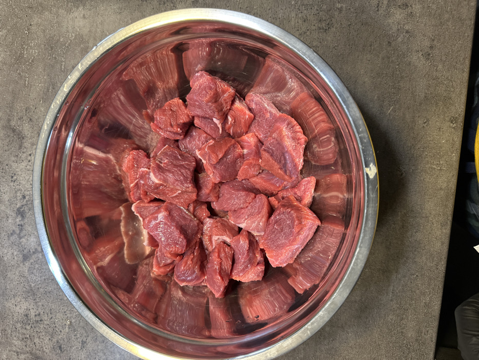 A large, round, reflective stainless steel mixing bowl, approximately 35-40 cm in diameter, is centrally positioned on a dark gray, textured surface resembling concrete or stone. The bowl contains numerous irregularly cut cubes of raw, lean red meat, ranging in size from approximately 2 to 5 cm per piece, exhibiting a fresh, reddish-pink color. The bowl's polished interior reflects the meat and surrounding light. Minor scuffs or wear marks are visible on the exterior rim of the bowl. A small portion of a dark-colored fabric or glove is visible at the extreme bottom-left corner of the frame, indicating the proximity of an individual. The scene is located in Tbilisi, Georgia, depicting a food preparation activity.