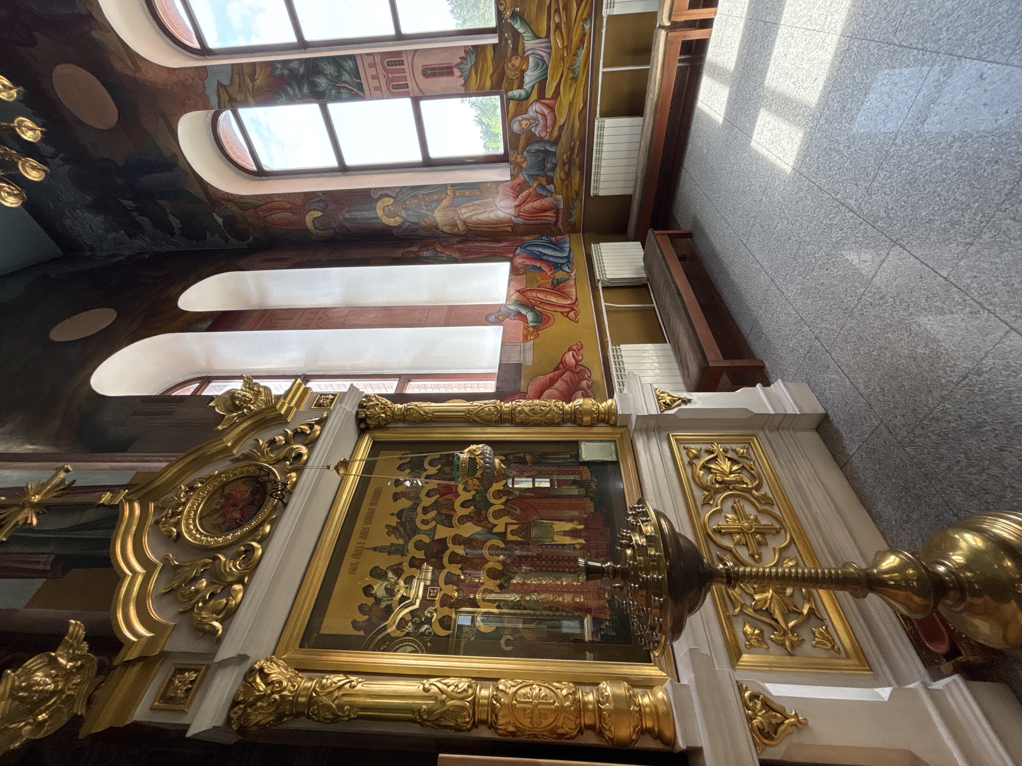 This interior view shows a decorated church space. A mural adorns the wall near the ceiling, depicting figures and structures. Windows are positioned near the top of the mural. The floor is tiled in a gray pattern. Ornate gold detailing surrounds a religious icon. A brass candlestick or lamp stands near the icon. The location is Chaikovsky, Russia.