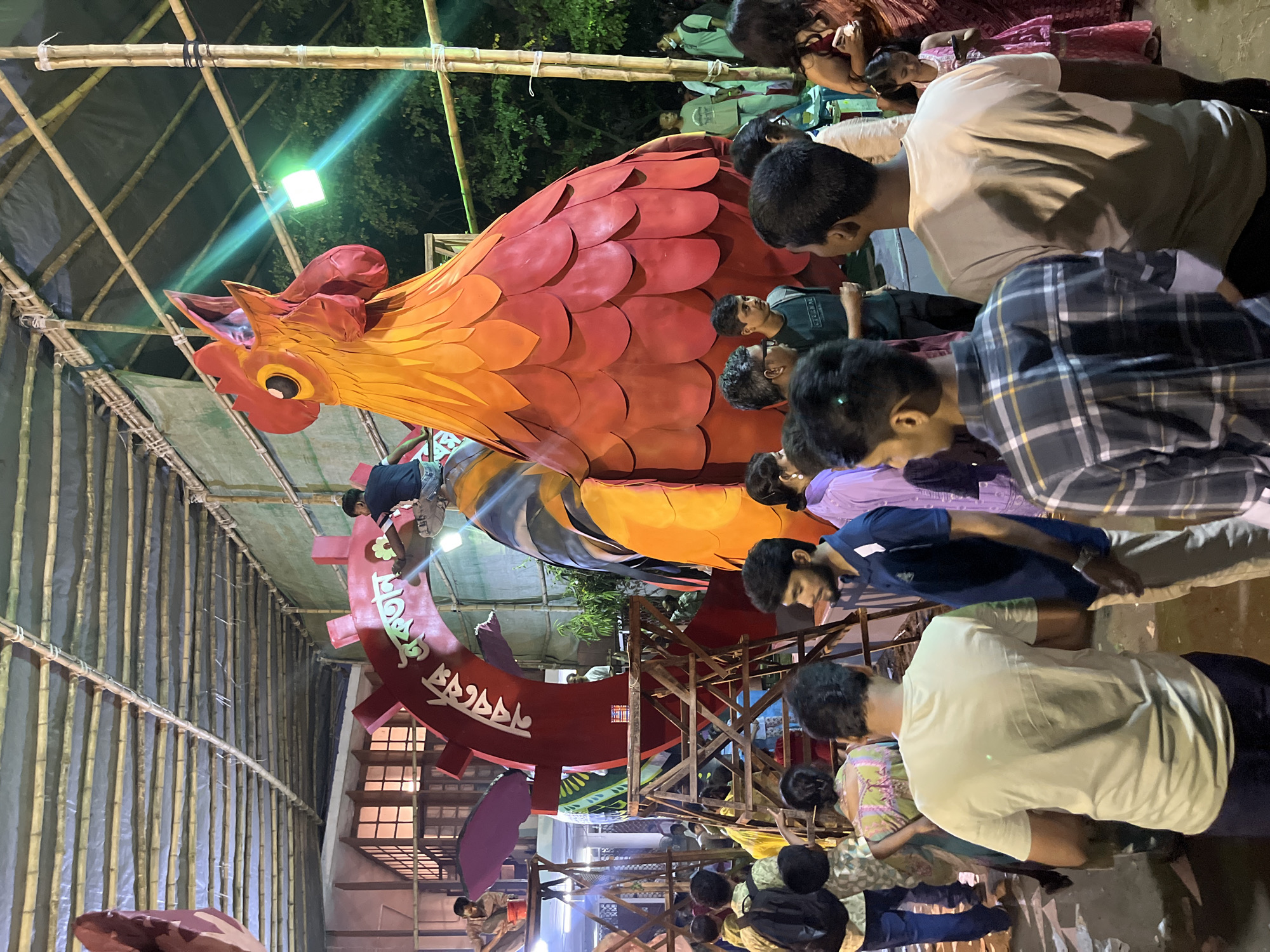 The photograph depicts a crowded street scene in Dhaka, Bangladesh, likely taken during a festival or public gathering. A large, elaborate rooster sculpture, predominantly red and orange, is the focal point. The rooster is constructed from multiple layers of overlapping elements, possibly representing feathers. It appears to be an art installation or part of a cultural display.
The surrounding environment is characterized by bamboo scaffolding and some other building construction. A mix of overhead lighting suggests it is nighttime or dusk.
Numerous people are present, mostly young men, standing close to the rooster sculpture. Most are looking at it. Some are casually dressed in shirts and trousers. The crowd suggests the event is popular and well-attended.
The scene conveys a sense of public engagement and cultural celebration. The specific location in Dhaka and the nature of the event cannot be determined with certainty, but it is evidently a significant gathering for the local community.
