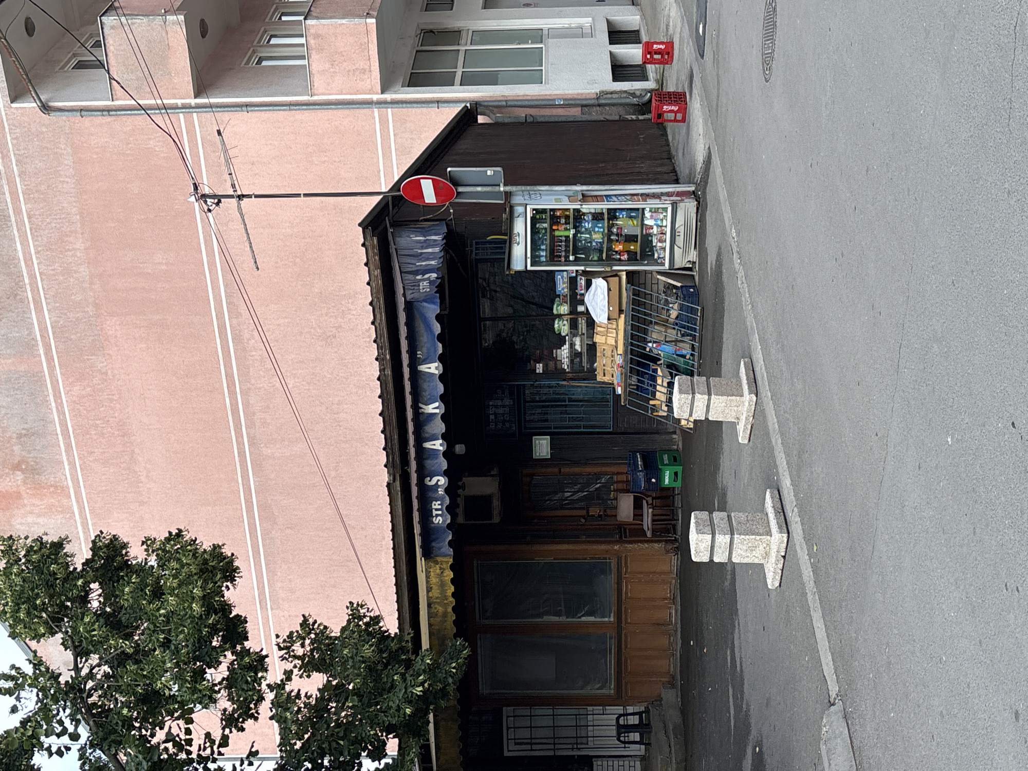 A street-level, exterior shot captures a small, rustic corner store with a blue awning that reads "STR S AKA." The store features wooden paneling and large glass windows, some of which are covered. In front of the store, a refrigerated display case is stocked with bottled beverages. A metal grate structure displays various goods, and crates of beverages are placed on the ground. A "no entry" traffic sign is mounted on a pole next to the store. The scene is situated on a paved street with concrete bollards lining the sidewalk. In the background, a pink building with white horizontal bands dominates the skyline, with a portion of a white modern building visible to the right. A large tree with green foliage partially obscures the top left of the image. The environment appears to be urban, with power lines and an antenna visible.