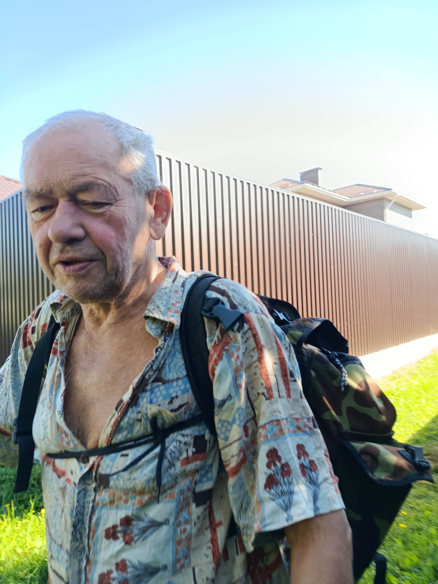 An elderly Caucasian man with white hair and a weathered face walks outdoors, carrying a camouflage backpack. He is wearing a colorful, short-sleeved shirt with a repeating pattern of red flowers and geometric shapes. The background consists of a tall, ribbed, dark brown fence, a section of a modern house with a tiled roof, and a bright blue sky. The ground in the foreground is covered with green grass. The scene appears to be a sunny day in Verea, Russia, possibly in a residential area.