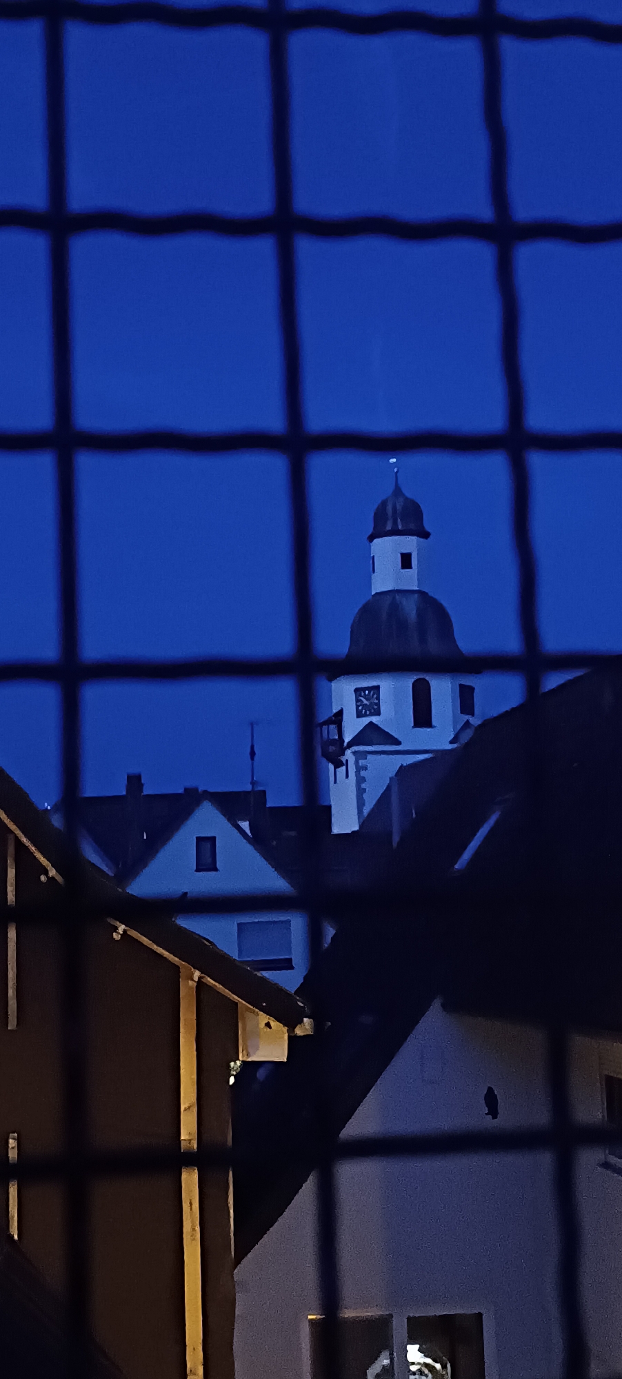 The image is a low-light, vertically oriented photograph taken at dusk or night. It depicts a view through a black metal grid, likely a fence or window covering, onto a cityscape. The sky is a deep, uniform blue, suggesting twilight.
In the background, a white church tower with a dark, domed roof and a spire is visible. The tower has a clock face with Roman numerals and a small balcony. Adjacent to the church, other buildings with dark roofs and illuminated windows can be seen. The foreground is dominated by the dark grid pattern.
Several residential buildings with pitched roofs and dark facades are also present, partially obscuring the view of the church tower and the sky. One building in the foreground has visible wooden structural elements on its exterior. The overall impression is of a residential area with a prominent church in the background, seen from a slightly elevated or obstructed vantage point.
The image was taken in Winnenden, Germany.