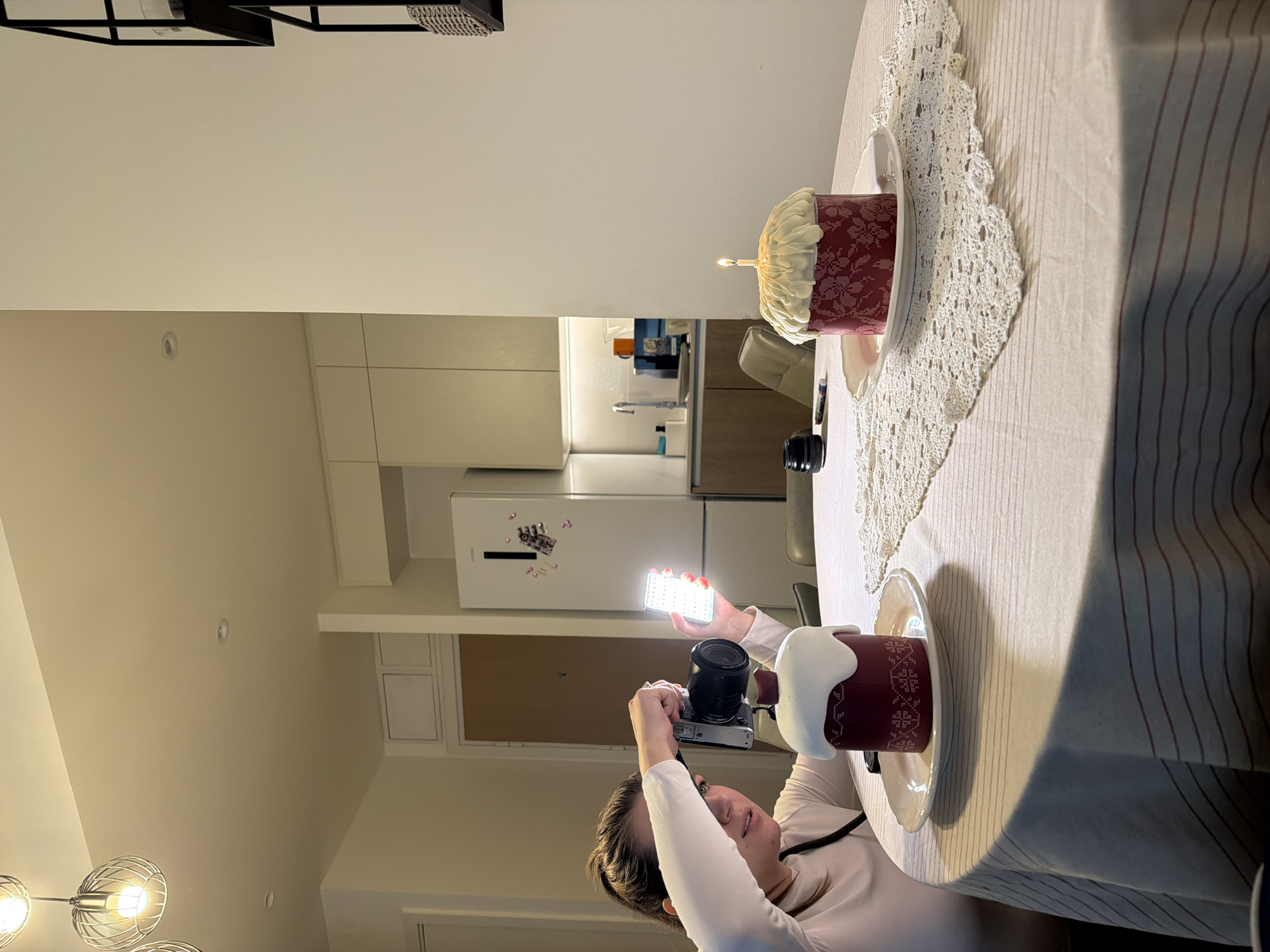 An indoor scene depicts a person photographing two cakes on a round table, situated within a modern apartment in what is identifiable as Belgrade, Serbia, given the Gorenje refrigerator and the traditional Orthodox Christian Easter bread (Kulich/Paska) cakes.
A female individual, viewed from the chest up, with her hair pulled back, is seated on the left side of a round table covered with a light-colored tablecloth featuring thin reddish horizontal stripes. She wears a long-sleeved light-colored top. Her left hand holds a silver-and-black mirrorless camera with a black lens, aiming it down towards a cake. Her right hand holds a rectangular LED panel light, illuminating the cake from the left.
On the table, there are two distinct cakes on individual white, fluted plates. The cake closer to the person is a burgundy-colored cylindrical cake, approximately 15-20 cm tall, adorned with white cross-stitch patterns. Its top is covered with a thick layer of white frosting, possibly melted, with a small burgundy element on top. A black camera lens cap rests next to this cake. Further to the right, another similar burgundy-colored cake with white cross-stitch patterns is present, also topped with white frosting, but this one features a single lit candle centrally placed on its crown, exhibiting a small flame. A white, crocheted square doily is spread beneath the second cake, overlapping the tablecloth. A black marker is visible near the second cake.
The background shows a contemporary apartment interior. Directly behind the person is a light-colored closed door. To the right, a white Gorenje refrigerator is visible, adorned with several small magnets. Beyond the refrigerator, a modern kitchen area is discernible with light-colored, handleless upper cabinets and darker lower cabinets. A sink with a chrome faucet and various kitchen items, including an orange mug and a blue container, are on the counter under task lighting. A light-colored chair is partially visible near the kitchen counter. Above, the ceiling is white with recessed lighting fixtures and two modern hanging light fixtures with exposed bulbs encased in wire cages. A tall, black, open-frame shelf unit is partially visible on the far right. The walls are painted a light, neutral color.