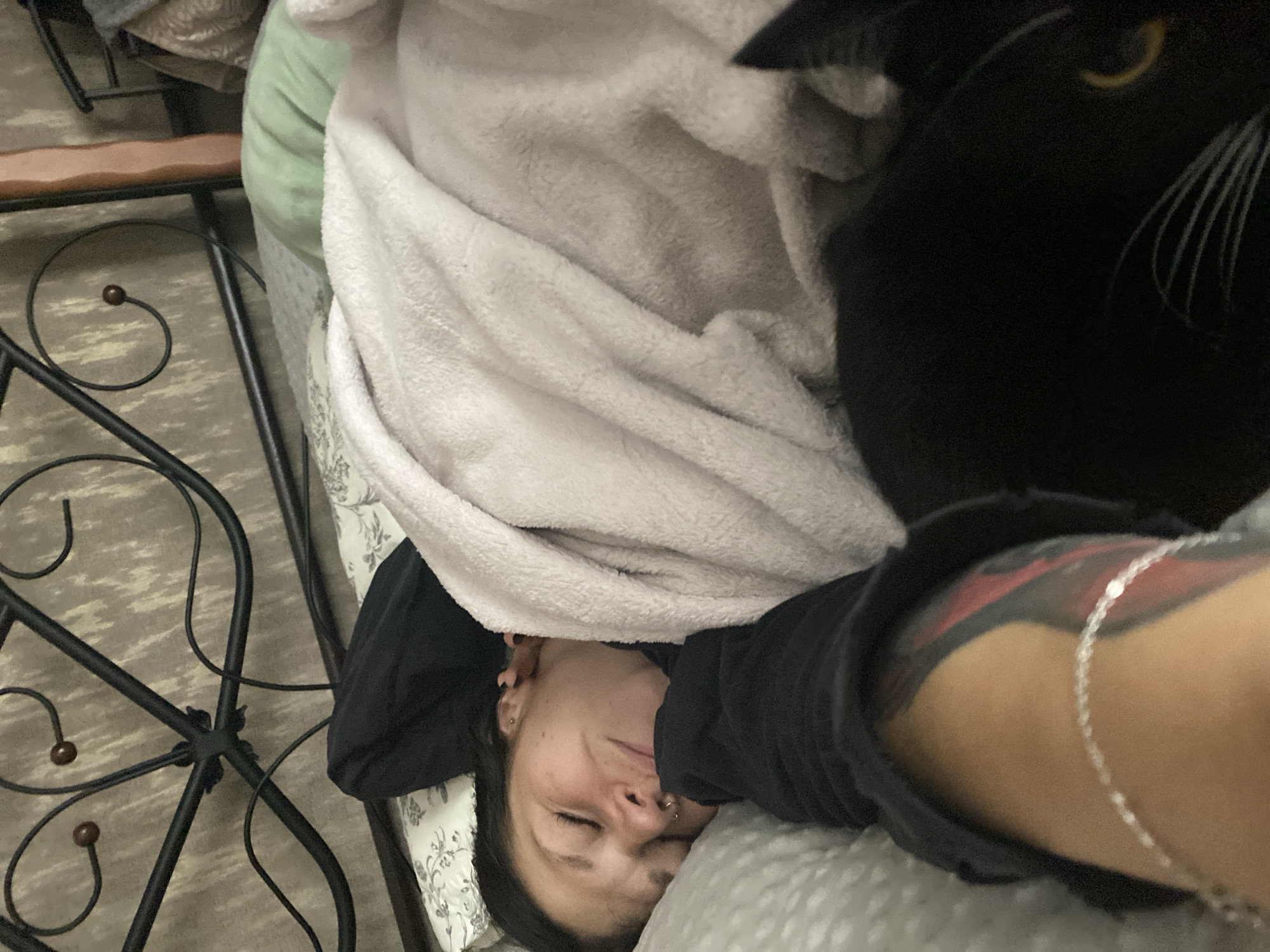 The image depicts a woman sleeping in bed, with a black cat nestled beside her. The woman is lying on her left side, her face turned towards the camera, with her eyes closed and a serene expression. She is covered by a light-colored blanket, and a black hoodie is partially visible. A silver anklet adorns her left leg, which also features a tattoo. The black cat is positioned to her right, its body largely obscured by the blanket and its head visible in the lower right corner, with one yellow eye peeking out. The background includes a metal bed frame and a textured wallpaper. The scene suggests a domestic setting, likely a bedroom, on a comfortable day. The photograph was taken in Chaikovsky, Russia.