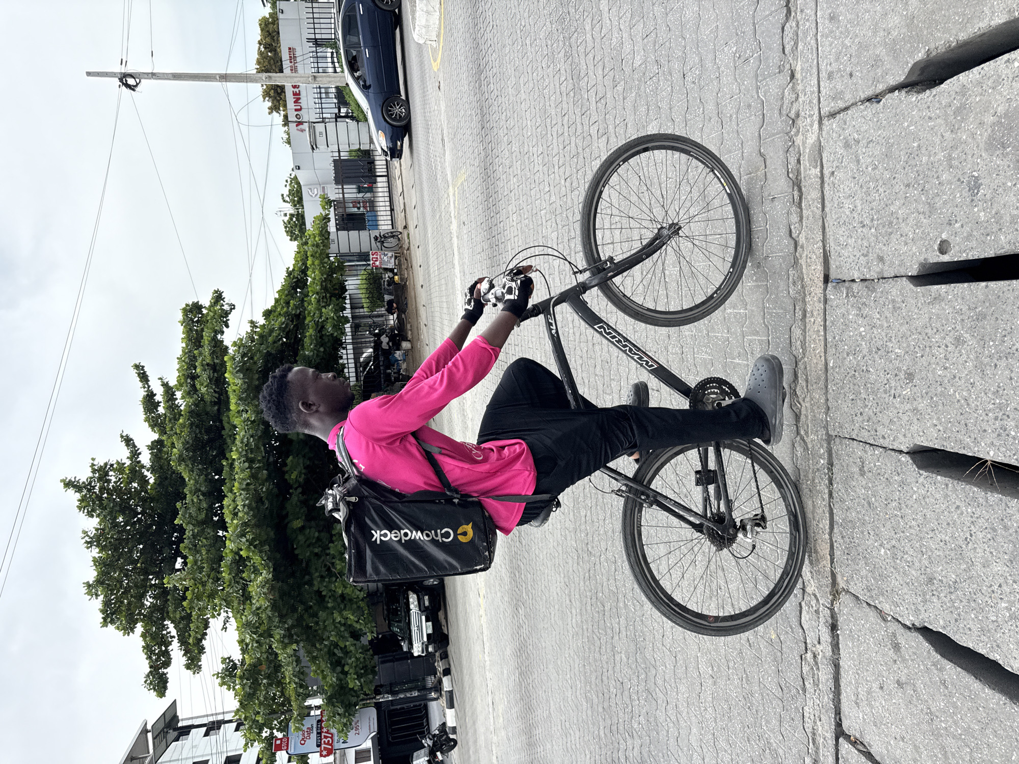 A man wearing a pink long-sleeved shirt, black pants, gray shoes, and black gloves is seated on a black Marin bicycle on a paved street. He is carrying a black "Chowdeck" delivery bag on his back. The street is adjacent to a curb with significant cracks.
In the background, there are buildings, trees, a dark blue car, and a utility pole with wires. Signs for GTCO, Quic Data, "JUST DIAL *737*", YOUNES, and POS are visible. The scene appears to be in Ikoyi, Nigeria, based on the signage.