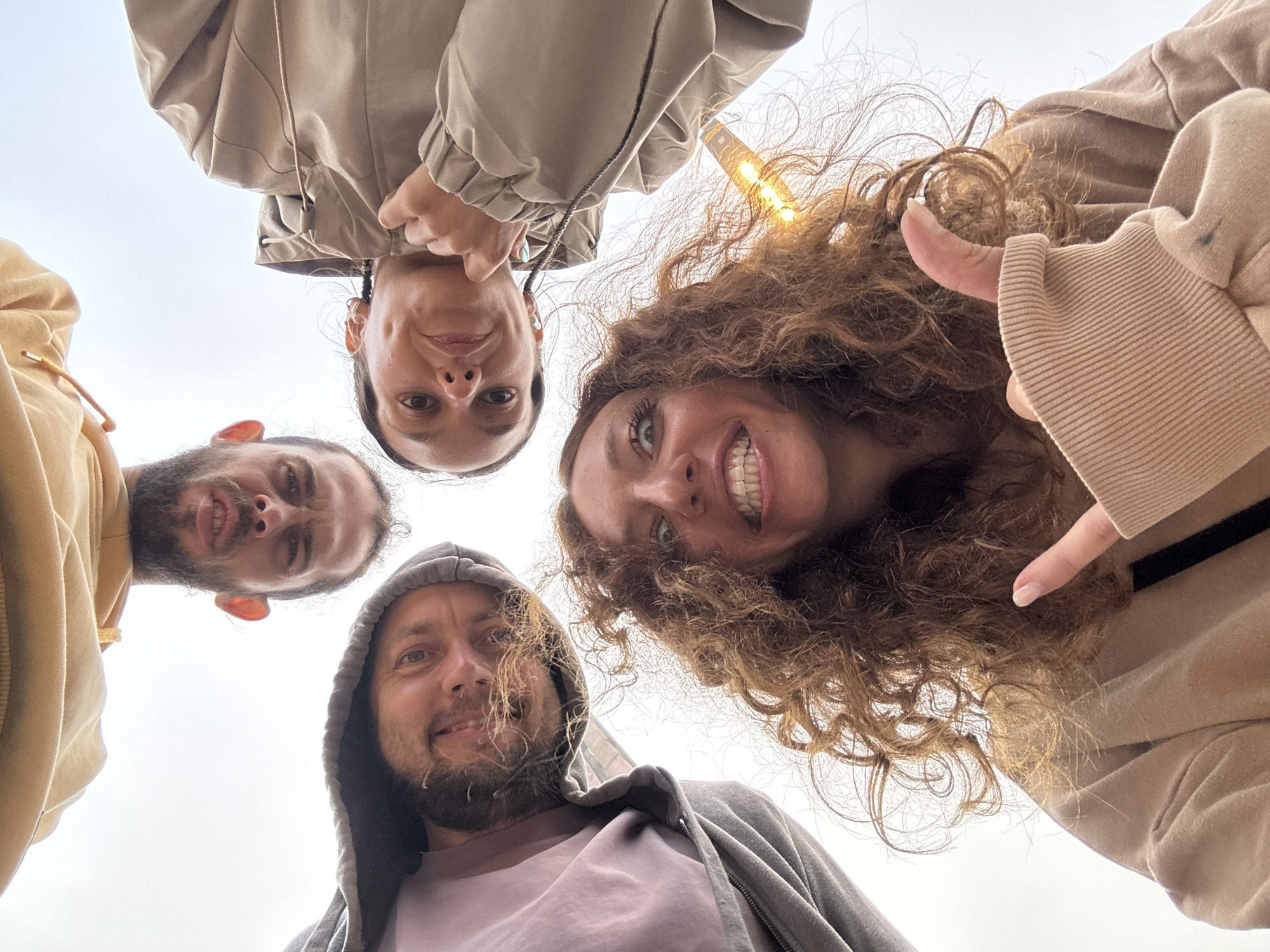 Four people are visible in the frame, arranged in a close-knit circle looking down at the camera. Two men have beards. Two people wear hoodies. The background is a cloudy sky and parts of buildings. The location is Binh Lu, Vietnam.