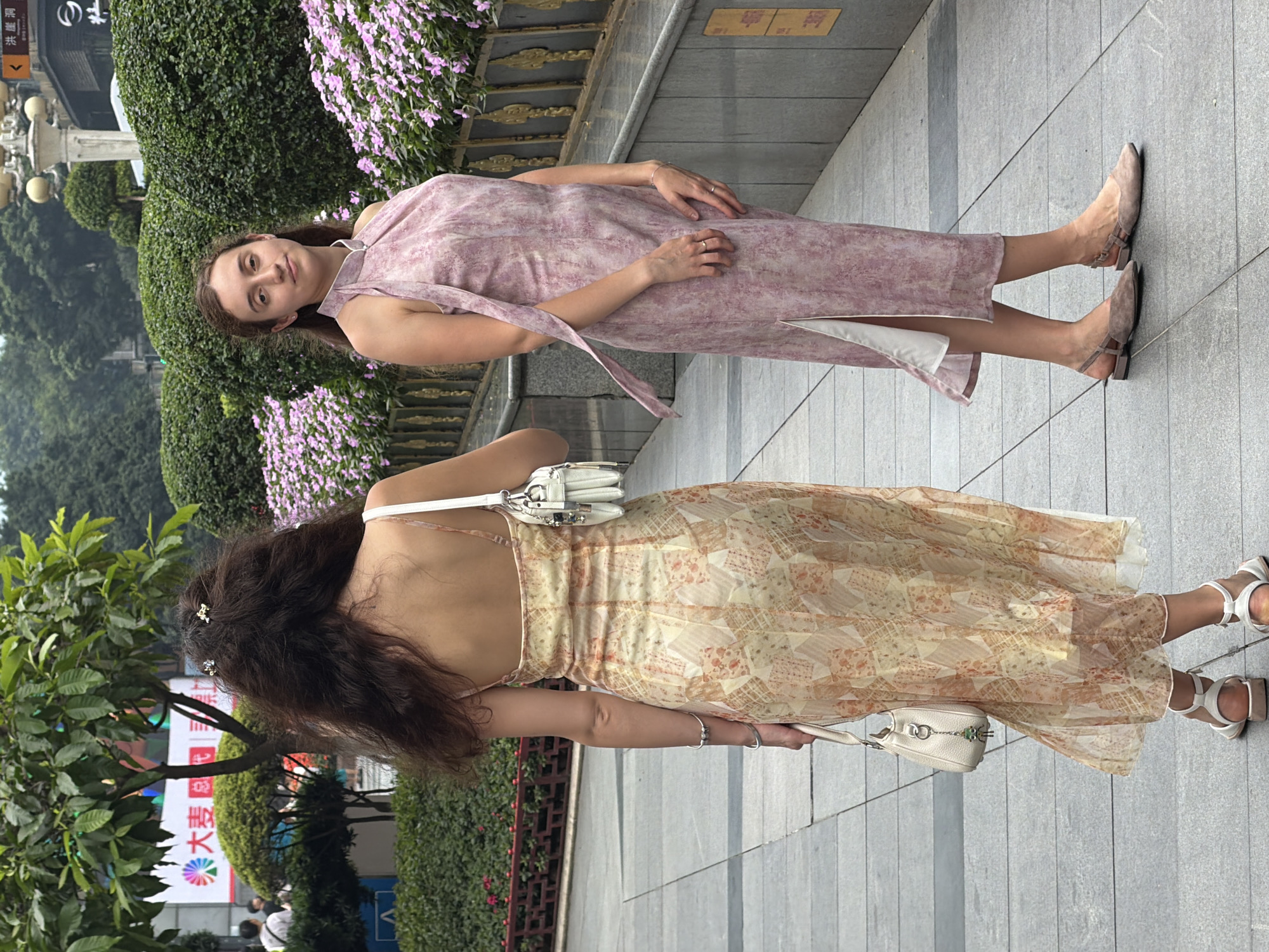 Two women are walking on a paved walkway in Yuzhong District, China. The woman on the left is viewed from behind, wearing a light yellow and orange floral print slip dress with spaghetti straps and white flat sandals. She carries a white handbag with a small green charm. The woman on the right is facing forward, wearing a pink floral print halter dress with a high neckline and a side slit, and beige slingback flats. She is positioned slightly behind and to the right of the first woman. Both women have jewelry on their hands. The background features lush green bushes, pink flowers in planters, a street lamp, and buildings. The lighting suggests an overcast day.