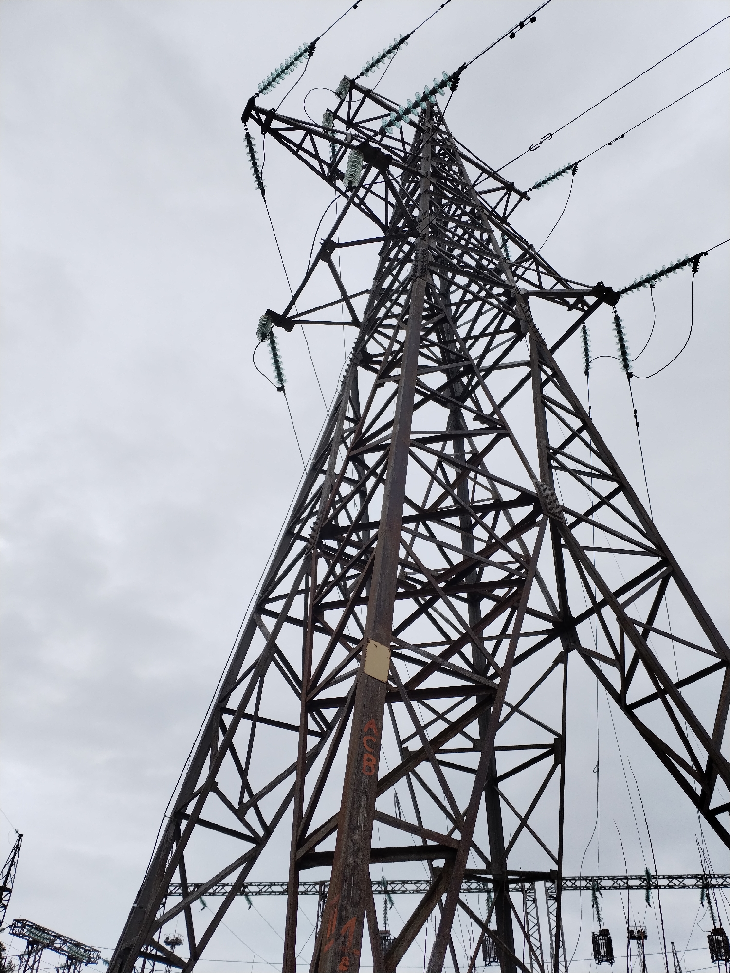 The image shows a steel lattice electricity pylon in Noviy, Russia, viewed from a low angle. The sky is overcast. The pylon structure is made of riveted steel beams forming triangular patterns. Electrical insulators are visible at the top of the pylon and along the support wires. Lower down, there is a small sign attached to one of the pylon legs.