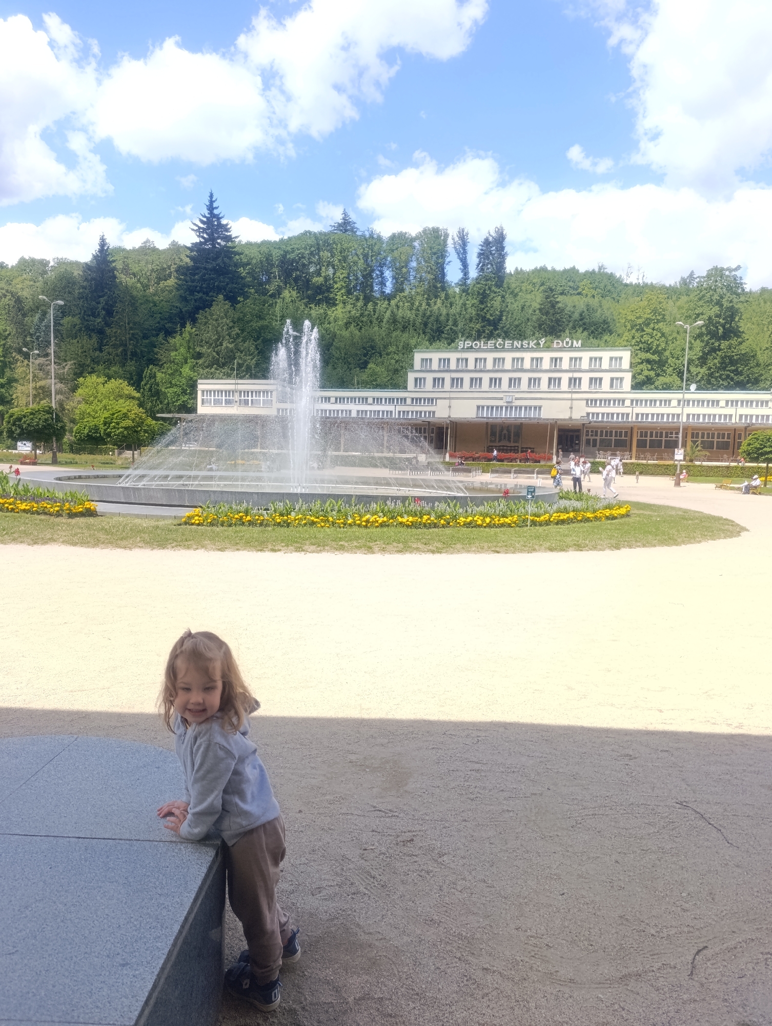 A young child with blonde hair is leaning on a grey stone bench in the foreground of the image, looking at the camera with a smile. The child is wearing a grey hoodie and brown pants. Behind the child is a large fountain spraying water, surrounded by a flower bed with yellow flowers. In the background, a long, light-colored building with many windows is visible, with text above reading "SPOLOČENSKÝ DŮM". The building is situated at the foot of a lush green hill covered in trees. The sky above is blue with scattered white clouds. This scene is located in Luhačovice, Czechia, a spa town known for its architecture and natural springs.