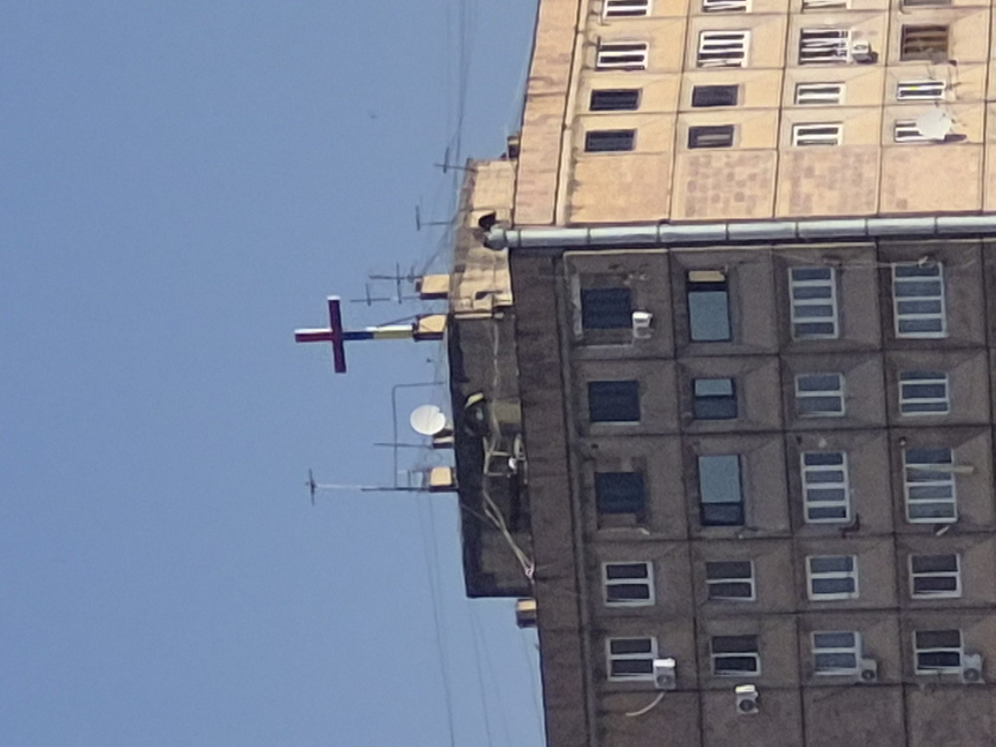 This vertical shot captures a section of a tall building against a clear, bright blue sky in Yerevan, Armenia. The building appears to be a multi-story structure, possibly residential or commercial, with a grid-like facade. It is composed of two main sections: an upper, lighter beige/brown part with square windows and a lower, darker grey-brown section also featuring numerous windows. A distinct horizontal band separates these two architectural styles.
A prominent feature is a colorful, cross-like structure extending horizontally from the side of the upper building section into the sky. This object is segmented with red, blue, and yellow colors, suggesting it might be a decorative element, a weather vane, or a flag-like display. Several antennas and satellite dishes are visible on the rooftop and various points along the building's exterior, indicating communication infrastructure. Numerous air conditioning units are also attached to the building's outer walls, particularly on the darker lower section.
The time of day is daytime, evidenced by the bright, clear blue sky and the crisp lighting on the building surfaces. The weather appears sunny with no clouds. No human activity is discernable, and there is no visible text in the image. The overall impression is one of an urban environment with utilitarian architecture.