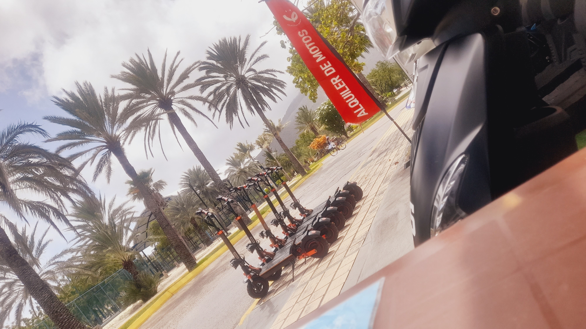 A row of electric scooters is parked on a paved walkway lined with palm trees. The scooters are black and orange, with a modern design. A red banner with white lettering that reads "ALQUILER DE MOTOS" (Motorcycle Rental) is visible in the background, suggesting a rental business. In the distance, mountains can be seen, and there is a person riding a bicycle. The sky is overcast, indicating a cloudy day. The scene appears to be in a tropical or subtropical location, consistent with the palm trees and the potential for scooter rentals. The overall mood is one of leisure and outdoor activity.