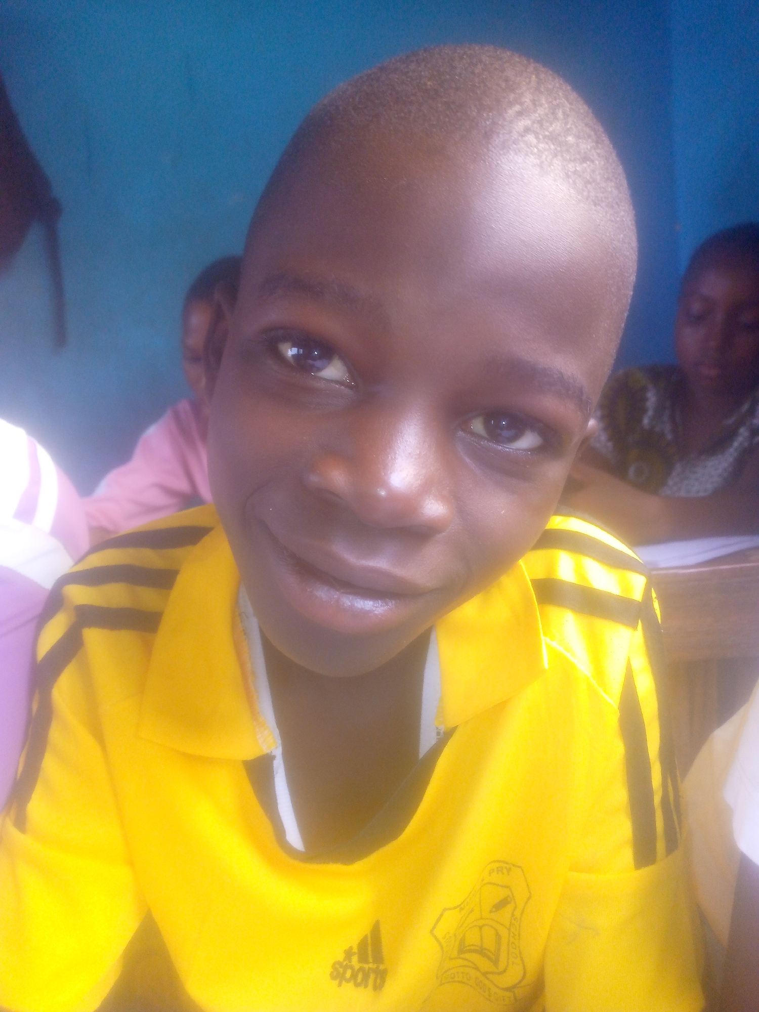 A close-up shot depicts a dark-skinned boy with a shaved head wearing a yellow Adidas t-shirt with black stripes on the sleeves. The boy is smiling and looking directly at the camera. Other children are blurred in the background, suggesting the scene is in a classroom. A turquoise-colored wall is visible in the upper portion of the frame. The location appears to be in the city of Unknown, Unknown.