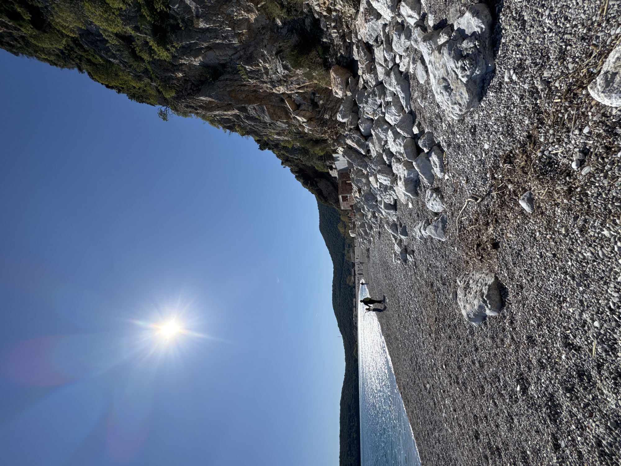 A bright, sunny day illuminates a scene at Prijevor, Montenegro. A long, narrow, pebble beach runs alongside the water's edge. Two people are visible on the beach. A rugged cliff face rises sharply on one side, its base fortified with large stones and rocks. The water shimmers under the sunlight.