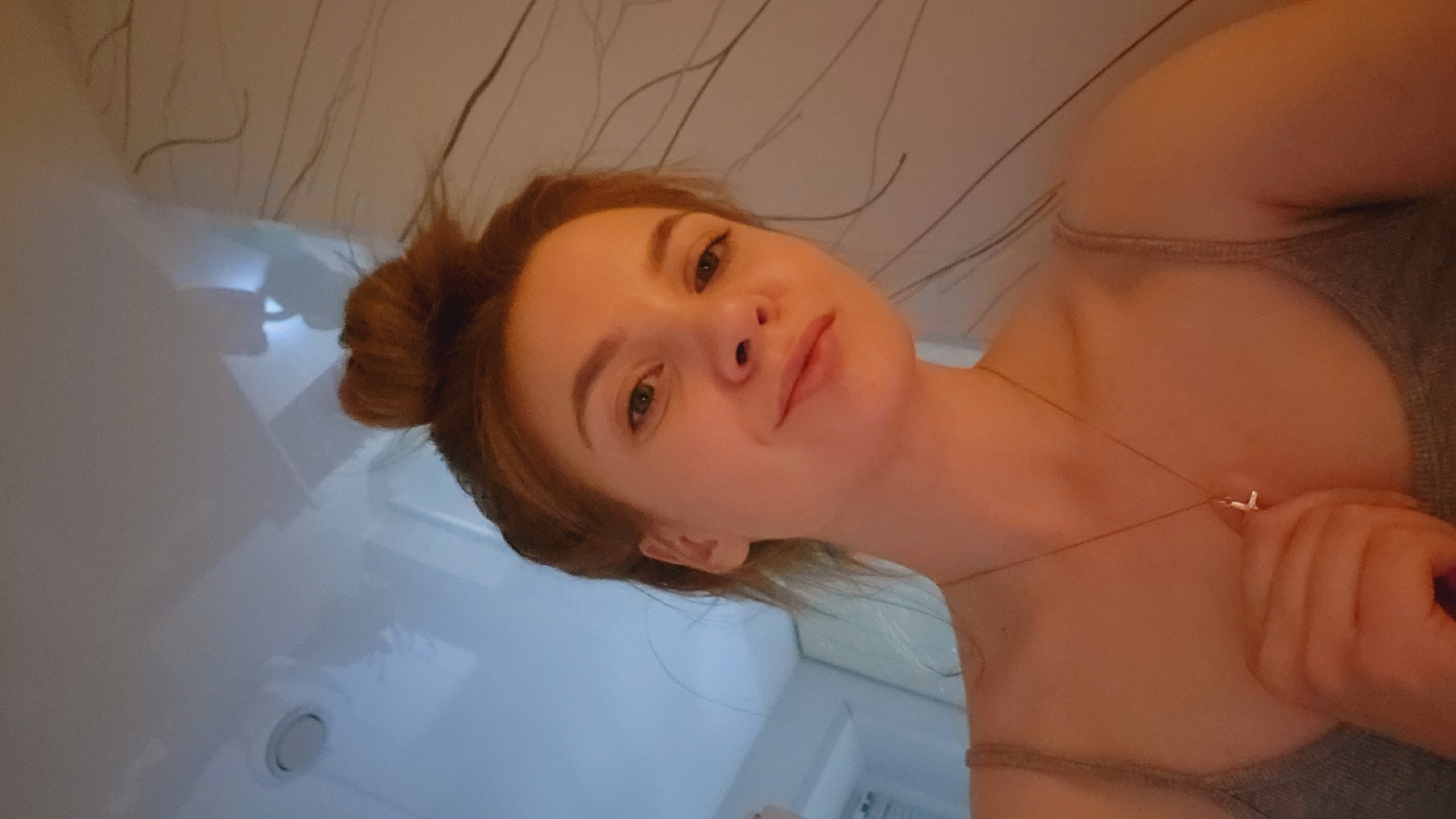 A medium shot captures a young woman in what appears to be a bathroom setting. She is angled, looking down at the camera, her face prominent. Her hair is gathered in a bun on top of her head. She is wearing a grey tank top and a delicate necklace.
Behind her, elements of a bathroom are visible. There's a white, possibly tiled, wall and a portion of what seems to be a shower or tub area. A round ventilation opening is visible on the wall.
Given the mention of Chaikovsky, Russia, this image would have been taken in an enclosed area within that city.