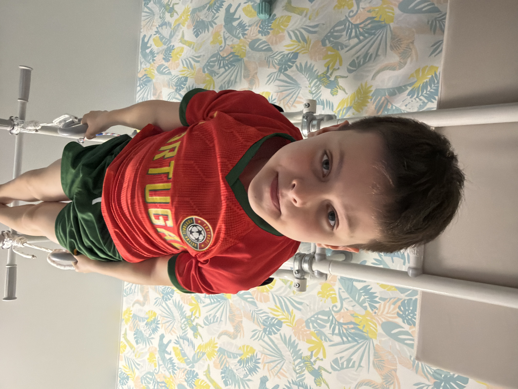 A portrait-oriented image, rotated 90 degrees clockwise, captures a young boy indoors, likely in a room in Votkinsk, Russia. The boy, appearing to be around elementary school age, has short dark hair and bright blue eyes, and is looking directly at the camera with a soft, friendly smile. He is dressed in a red short-sleeved soccer jersey with green trim, featuring "PORTUGAL" in yellow lettering on the front, and a circular soccer team crest. He also wears dark green shorts.
The boy is interacting with a white indoor play or fitness structure, likely a Swedish ladder or similar climbing frame, holding onto vertical poles equipped with gray handles. The background behind him showcases a lively wallpaper pattern, predominantly in light blue, yellow, and peach tones, depicting tropical leaves and subtle animal figures that resemble lizards or chameleons. The bright, diffuse lighting suggests an indoor daytime setting. The scene portrays the child in a relaxed and playful manner within his home environment.