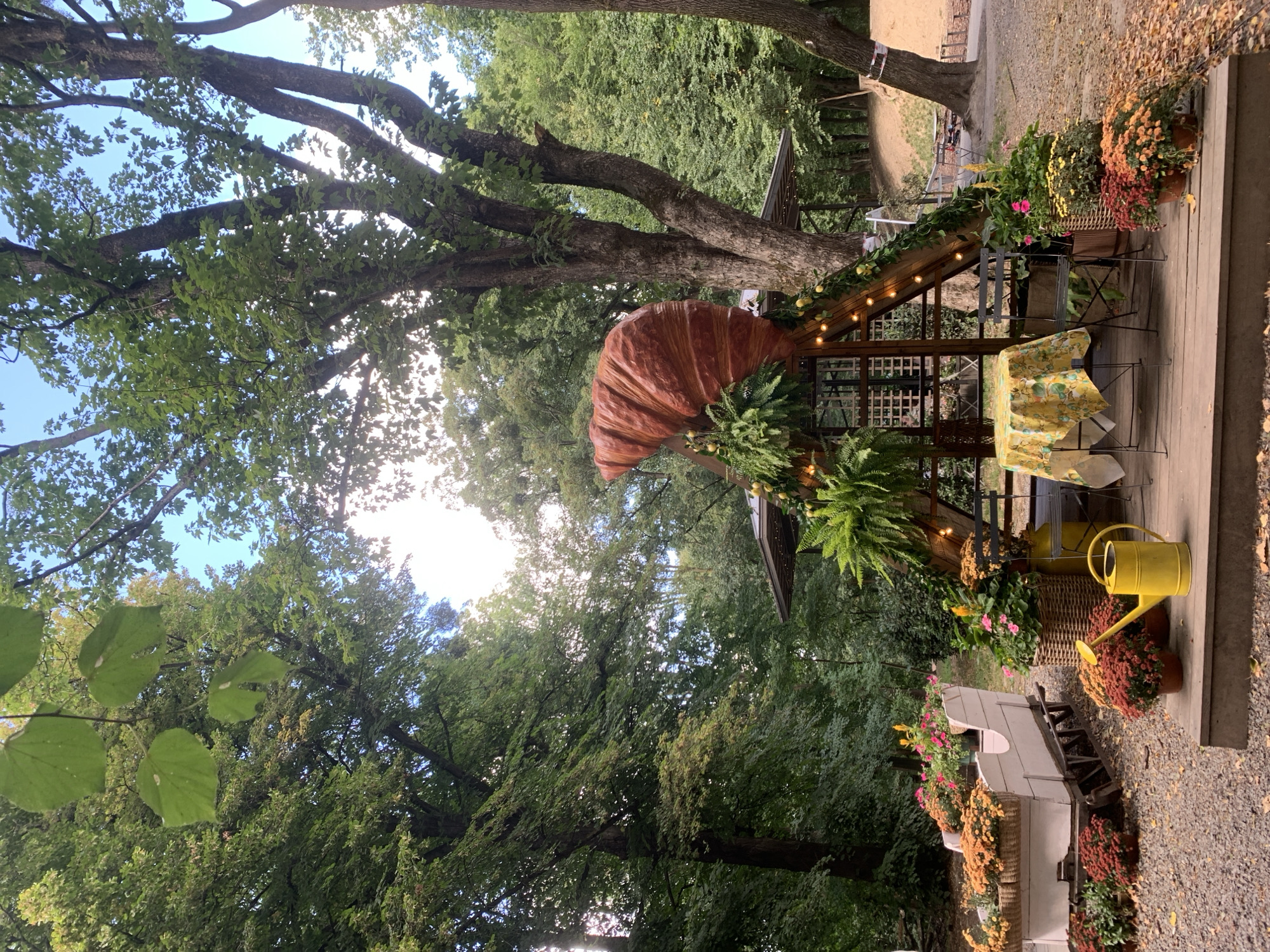 The image depicts an outdoor scene in Lviv, Ukraine, characterized by a rustic, nature-inspired aesthetic. In the foreground, a wooden deck is furnished with a small table covered with a yellow and green patterned tablecloth, accompanied by two dark metal chairs. Surrounding the deck are potted chrysanthemum flowers in various shades of red, orange, and yellow, along with a bright yellow watering can. To the left of the deck, a white, sleigh-like planter overflows with more orange and yellow flowers.
Dominating the center of the scene is a wooden structure adorned with greenery and string lights. Perched atop this structure is an oversized, realistic croissant sculpture. The entire setup is set against a backdrop of lush green trees, suggesting a park or garden environment. The ground is covered with gravel and fallen leaves, further contributing to the autumnal atmosphere. The sky above is visible through the tree canopy, appearing partly cloudy. The overall impression is of a charming, possibly seasonal, outdoor seating or display area.