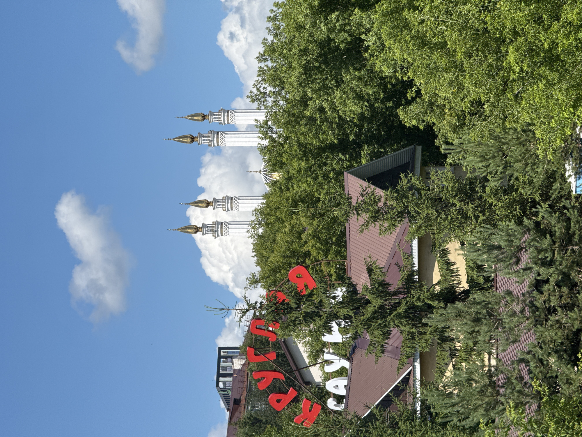 An outdoor, eye-level, medium shot captures a collection of structures and foliage under a bright, blue, sky with scattered white clouds. In the background, four white minarets with golden spires pierce the sky, suggesting a religious or historical site. A partially obscured golden dome is also visible behind lush green trees. In the mid-ground, a large, bright red sign with Cyrillic lettering, possibly advertising a business, is mounted on a stand amidst the trees. Below this sign, a white sign with more Cyrillic lettering is visible. The foreground is dominated by the dark brown, sloped roof of a building, with several evergreen trees in front of it. The overall scene is one of a partially urban, partially natural environment.