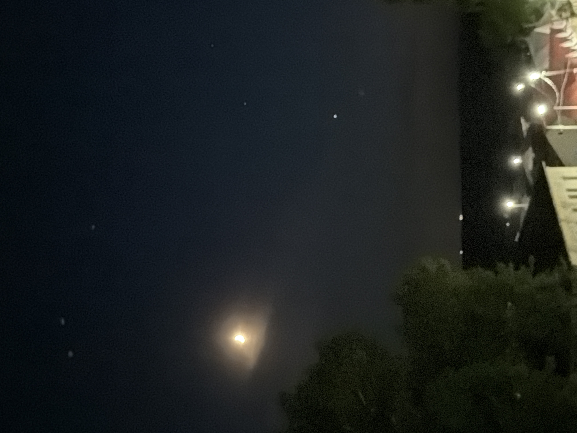 The image depicts a night scene, likely in Chaikovsky, Russia, though this is not definitively discernible from the image alone. The sky is dark, with a partially visible moon near the bottom center. A few faint stars are scattered across the sky. Trees are visible in the foreground on the right and bottom of the frame, and a structure with string lights is visible in the upper right corner. The scene is dimly lit. There are no people or actions apparent in the frame.