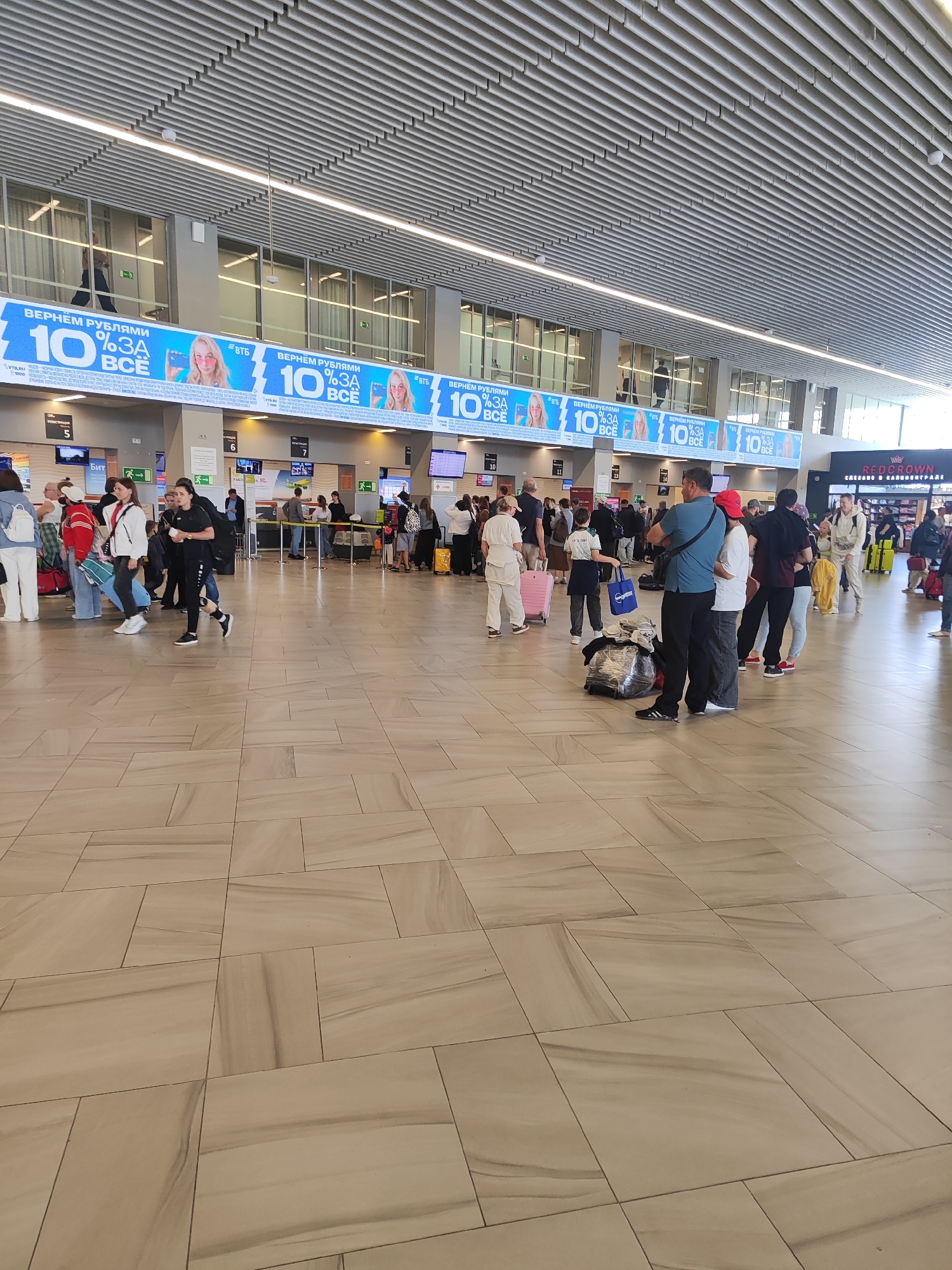 The image depicts the interior of an airport terminal in Khrabrovo, Russia. In the foreground, the floor is tiled with large, light-colored rectangular tiles with a marbled pattern. A crowd of people, appearing to be travelers, are scattered throughout the scene. They are standing, walking, and queuing, many with luggage.
In the middle ground, there are several check-in counters with digital displays and signage. The check-in counters are numbered from 5 to 14. Above the check-in counters, a large advertising banner is displayed across the width of the terminal. This banner features the text "ВЕРНЁМ РУБЛЯМИ 10% ЗА ВСЁ" (We'll return in rubles 10% for everything) and includes images of a woman with pink sunglasses and branding for "ВТБ" (VTB Bank).
Further in the background, more travelers and airport personnel are visible. There are also information screens and the general architecture of a modern airport terminal, including a high ceiling with linear lighting and ventilation systems. The overall atmosphere appears to be one of transit and activity associated with air travel.