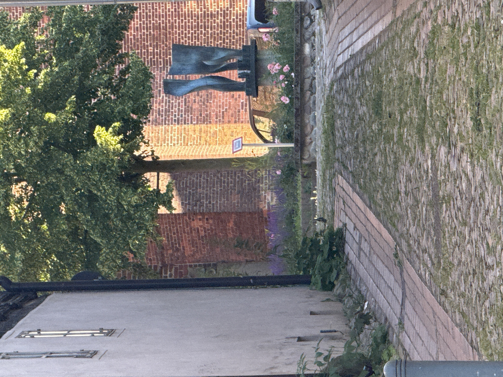 An outdoor, eye-level shot shows a narrow alleyway paved with cobblestones and bordered by a light-colored building on the left and a brick building on the right. The alleyway curves slightly to the right as it recedes into the distance. Green foliage grows along the base of the buildings, and a large tree with lush green leaves is visible in the background, casting shadows on the brick wall.
In the middle ground, a sculpture of two abstract, blue human legs stands on a stone pedestal. Pink roses and purple flowers bloom around the base of the sculpture, and a car with a reflective surface is parked behind it. The brick building to the right appears to be an old church or historical structure, indicated by its architecture and the presence of a small, red-and-white sign with an icon near the sculpture.
The lighting suggests it is either early morning or late afternoon, with warm sunlight illuminating the scene and creating long shadows. There are no people clearly visible in the foreground or mid-ground. The overall impression is one of a quiet, historic urban setting.