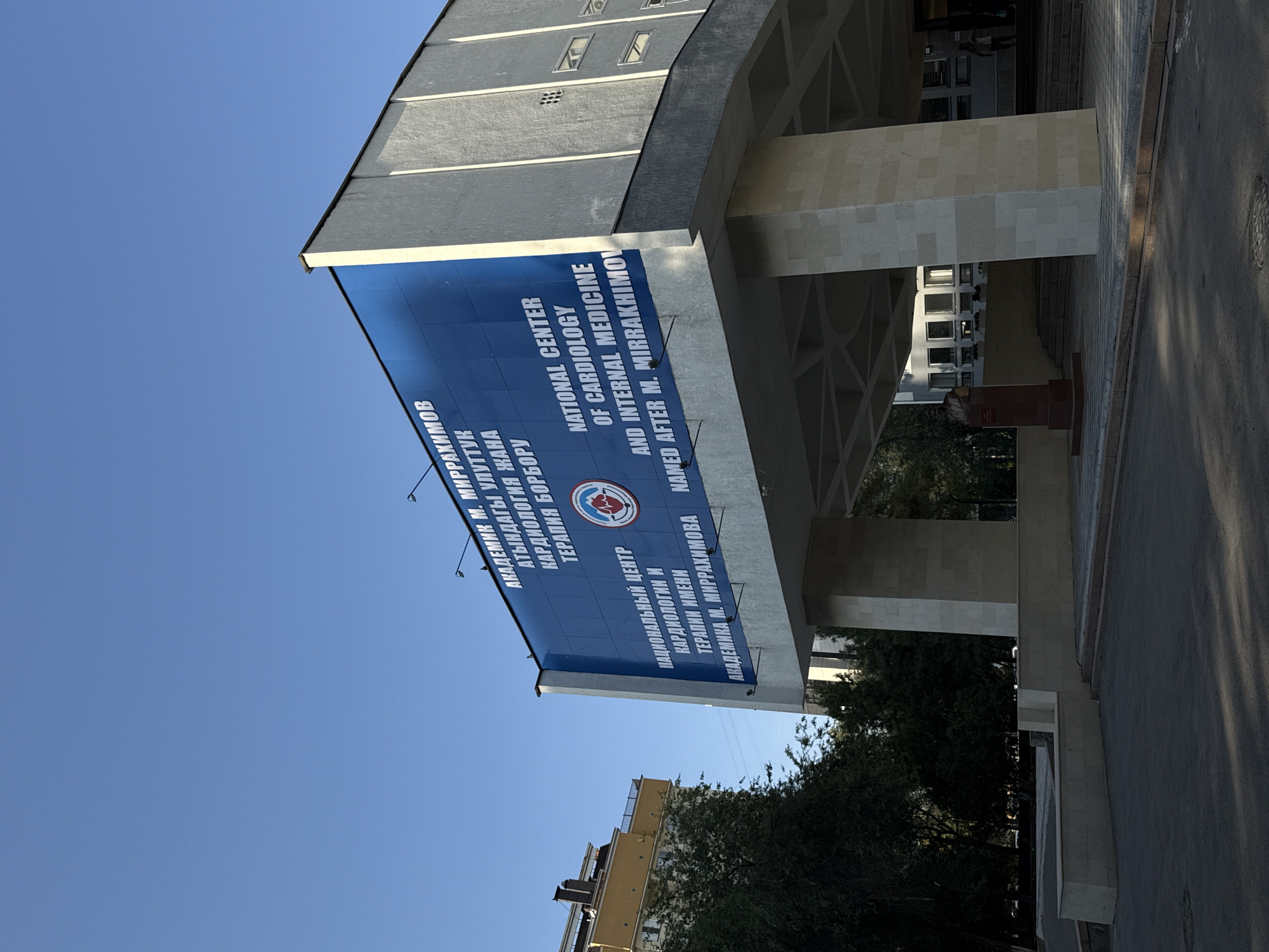 The image depicts the National Center of Cardiology and Internal Medicine named after M. Mirrakhimov in Bishkek City, Kyrgyzstan. A large blue banner with white text spans the upper facade of the building. The text is in both Russian and English, with the English text reading "NATIONAL CENTER OF CARDIOLOGY AND INTERNAL MEDICINE NAMED AFTER M. MIRRAKHIMOV." A circular logo with a heart and EKG line is also visible on the banner. The building itself is a modern concrete structure with large pillars supporting an overhang. In the foreground, a statue of a man is visible on a pedestal. The surrounding environment includes trees and a clear blue sky. The image appears to have been taken during the daytime.
