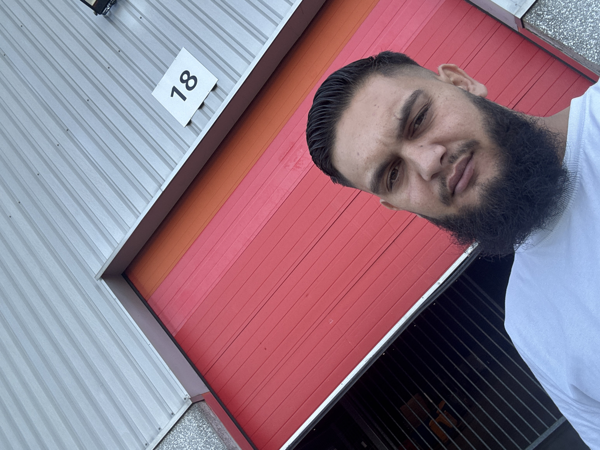 A man with a beard and short dark hair, wearing a white t-shirt, is taking a selfie in front of a large red garage door that is partially open, revealing a dark interior with horizontal shelving units. Above the red door, there is an orange section and then a corrugated gray metal roof. A white square sign with the number "18" in black is affixed to the gray roof. The scene appears to be an outdoor industrial or storage area. The location is identified as Boom, Belgium.