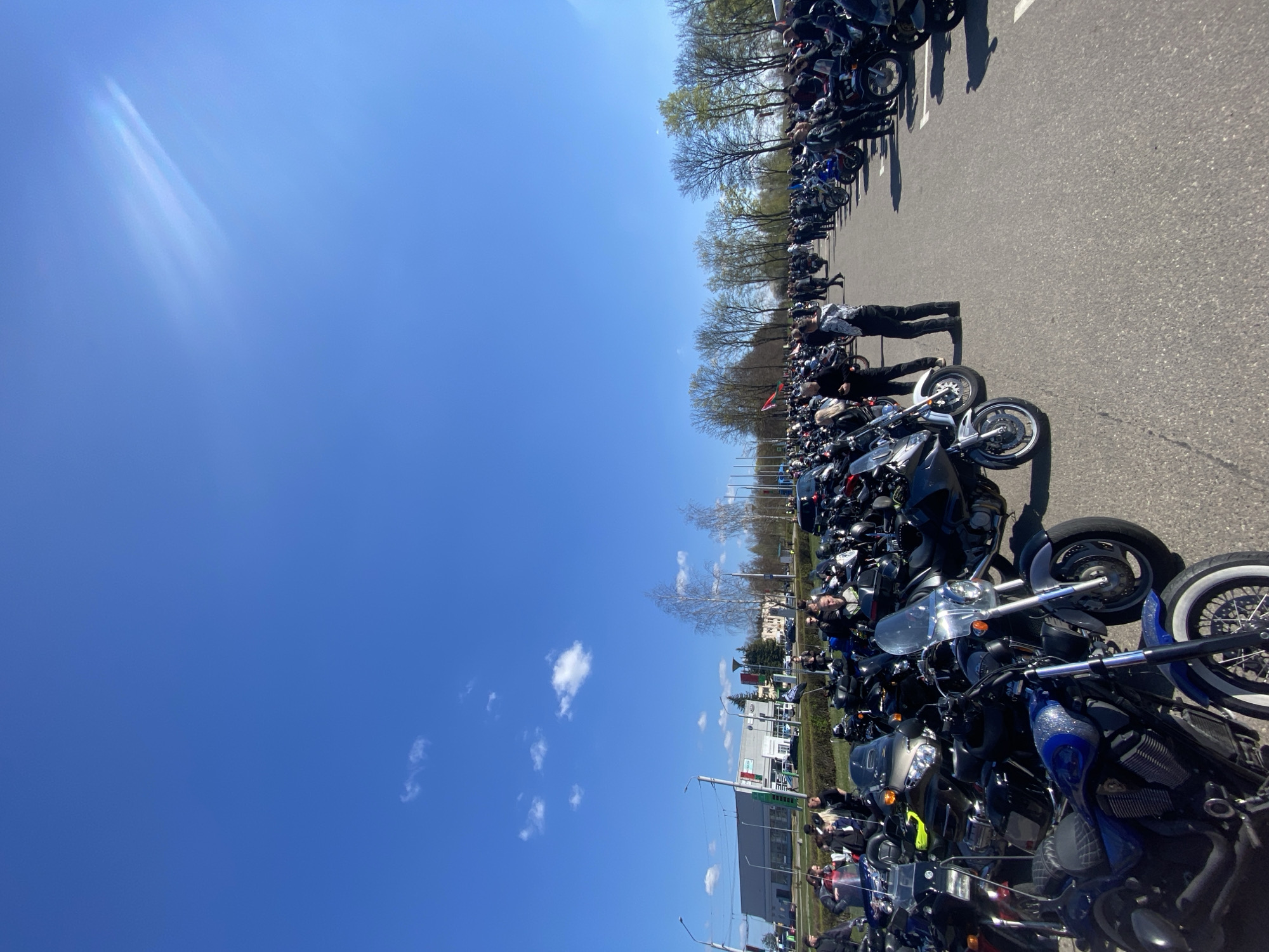 The image depicts a large gathering of motorcycles parked along a road in Hrodna, Belarus. The motorcycles are lined up in multiple rows, suggesting an organized event or rally. Individuals are standing and interacting among the bikes. Behind the motorcycles are trees and buildings. The sky is blue with scattered clouds.