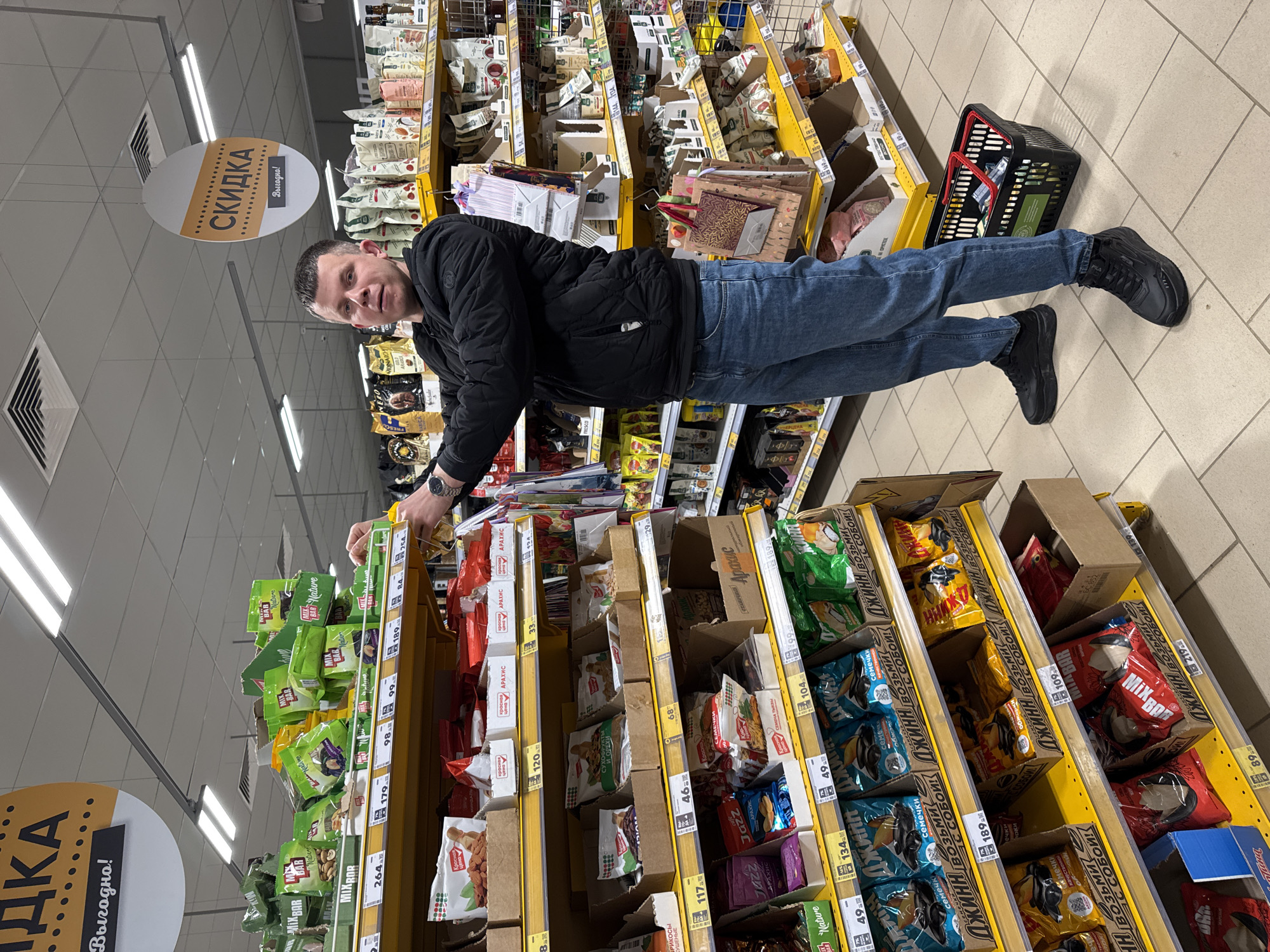 A mid-height shot captures a man standing in the aisle of a grocery store, presumably in the city of Горный, Russia. The man, dressed in a black jacket and blue jeans, is facing the camera while reaching towards a shelf filled with packaged snacks, likely nuts or dried fruits.
The store's interior is brightly lit, featuring rows of shelves packed with various goods. The aisle is well-stocked with neatly organized product displays. The shelves are marked with price tags in Russian currency. Signage indicating "Скидка Выгодно!" (Discount Profitable!) is visible in the upper left, emphasizing sales promotions.
The man's shopping basket is visible near his feet. The overall setting suggests a routine shopping trip.