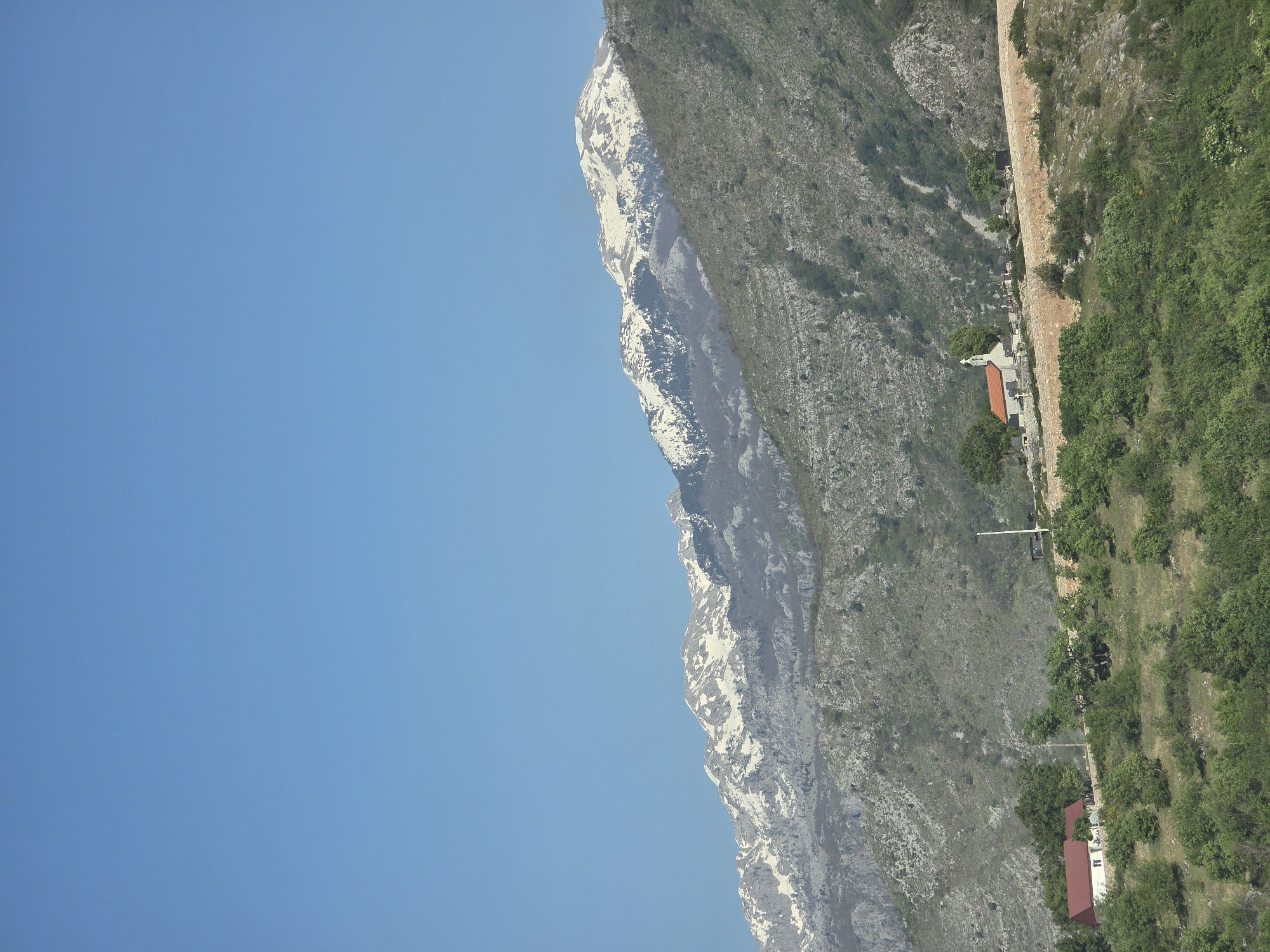 An eye-level, long shot shows mountains on the edge of Klopot, Montenegro, on a clear day. The left side of the image contains a clear, bright blue sky. From bottom to top on the right side is a mountain range covered with green foliage. The edge of the mountain range is snowy. At the bottom of the mountain range are buildings with red roofs. Beside the buildings is more green foliage.
