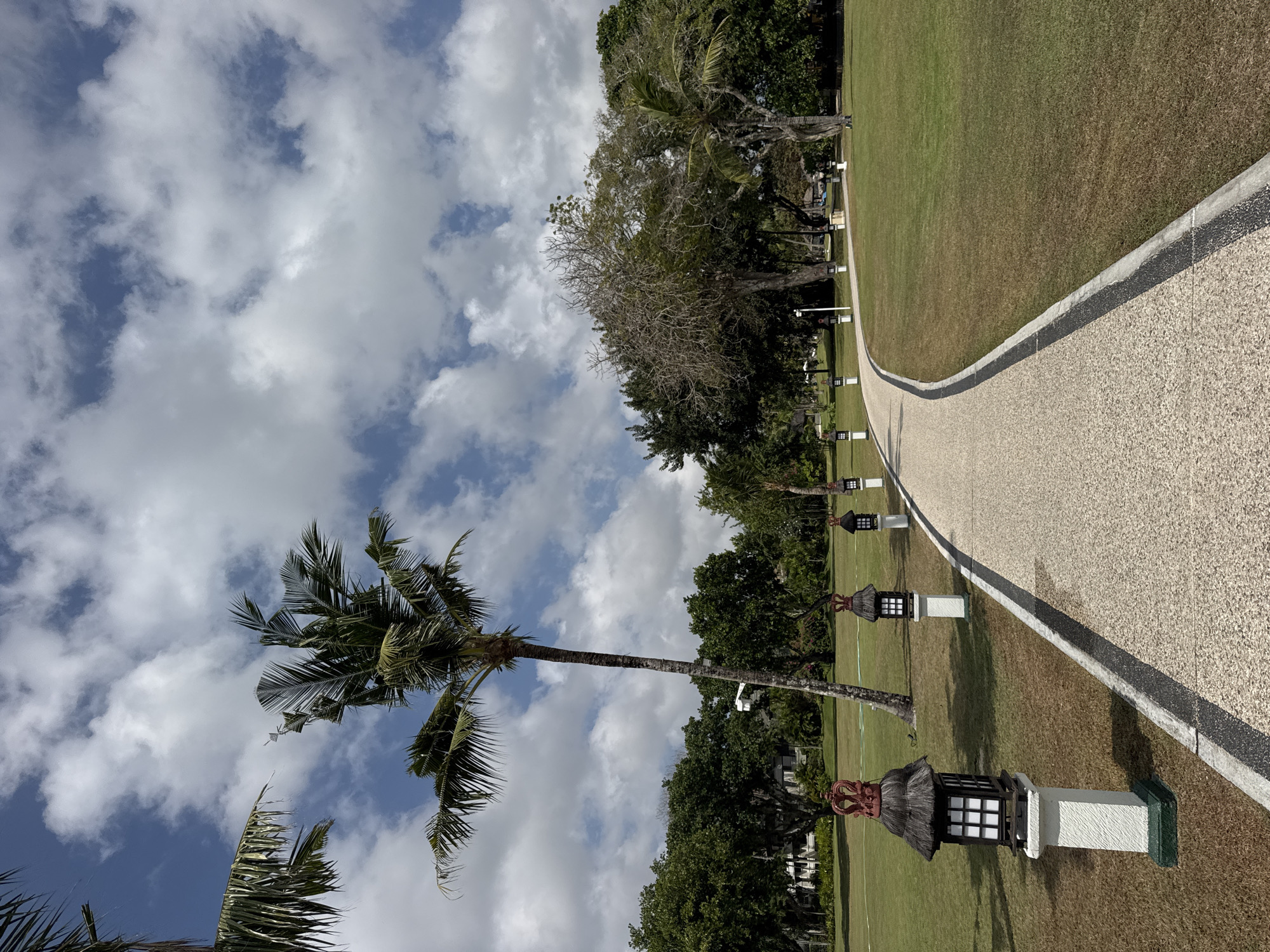 The image depicts a curved pathway made of light-colored gravel with a dark border, winding through a well-maintained lawn. Along the left side of the pathway, several ornate garden lamps with thatched roofs and white pedestals are visible. In the background, lush green trees and a tall palm tree are present, with a few buildings peeking through the foliage. The sky is blue with scattered white clouds, suggesting daytime. The scene appears to be a resort or a park area, likely in the city of Kuta Selatan, Indonesia, given the tropical vegetation and architectural style of the lamps. There are no people or animals visible in the image.