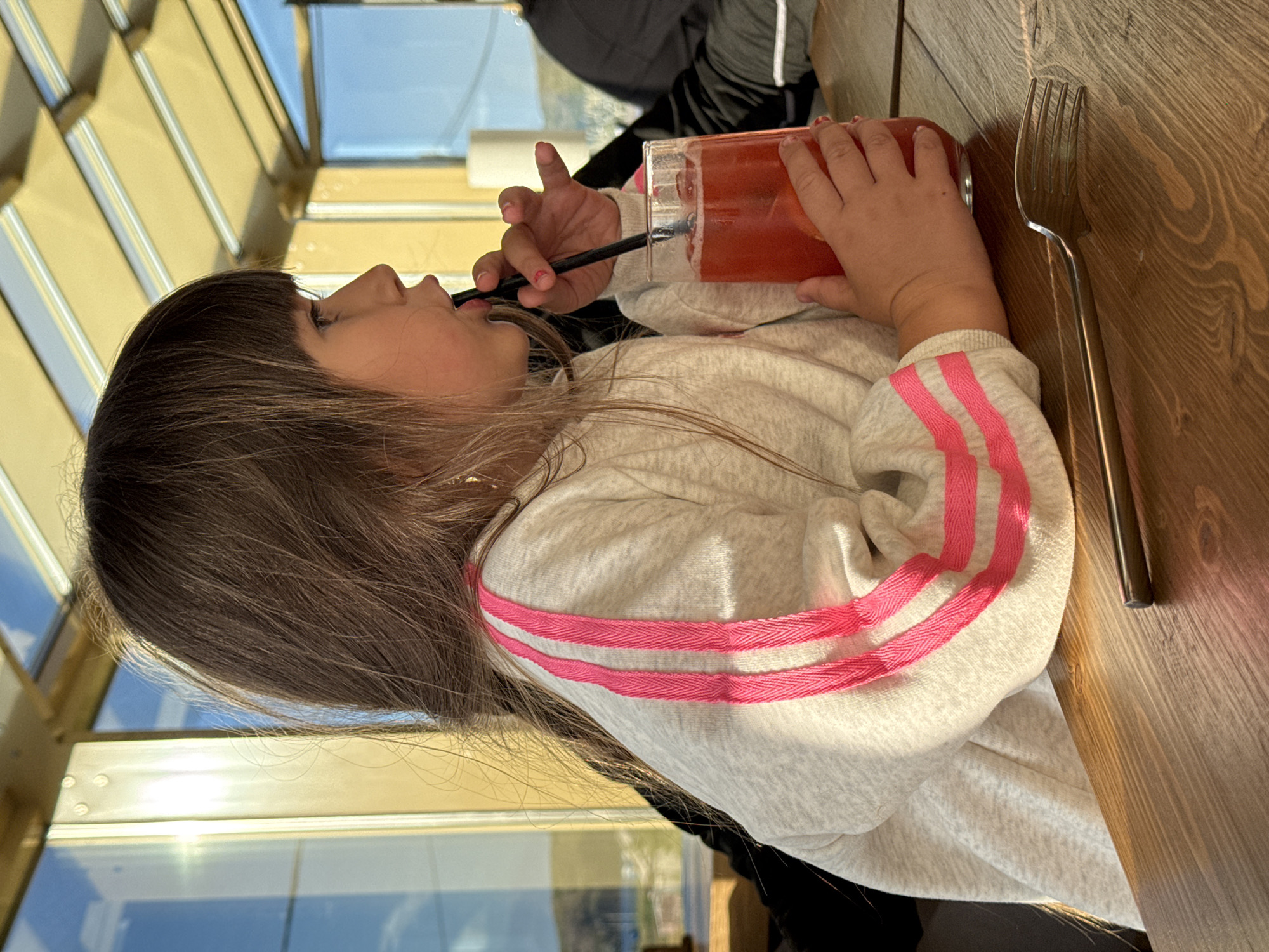 A young girl with long, brown hair and bangs is sitting at a wooden table, holding a glass of red liquid with ice and a straw. She is wearing a light gray sweatshirt with pink stripes on the sleeves. The girl is looking to her right and drinking from the straw. A fork is also on the table in front of her. The background shows large windows with sunlight streaming in, reflecting the sky and some buildings outside. The setting appears to be a restaurant or cafe, possibly in a location with a view of the water and hills, consistent with the coastal environment of Teriberka, Russia. The lighting suggests it is daytime.