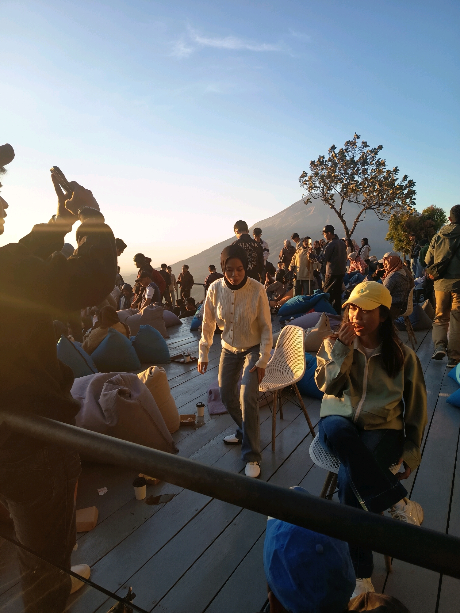 The image depicts an outdoor gathering at a viewpoint overlooking a mountain, likely Mount Semeru, near Keditan, Indonesia. The scene is illuminated by the soft light of sunrise or sunset. A wooden deck serves as the gathering space, furnished with numerous beanbag chairs and a few regular chairs.
In the foreground, a person is partially visible, holding a phone up to capture a photograph, with their arm silhouetted against the bright sky. To the right, a young woman wearing a yellow cap and a light olive jacket is seated, eating something and looking to her left. Another young woman in a cream-colored cardigan walks past her.
In the background, a diverse group of people are scattered across the deck, some standing and others sitting on beanbags or chairs. The mountain, possibly Mount Semeru, is visible in the distance, with a lone tree in the mid-ground framing it. The sky is a clear blue with a few wispy clouds. The overall atmosphere appears relaxed and social, with individuals enjoying the view and each other's company.
