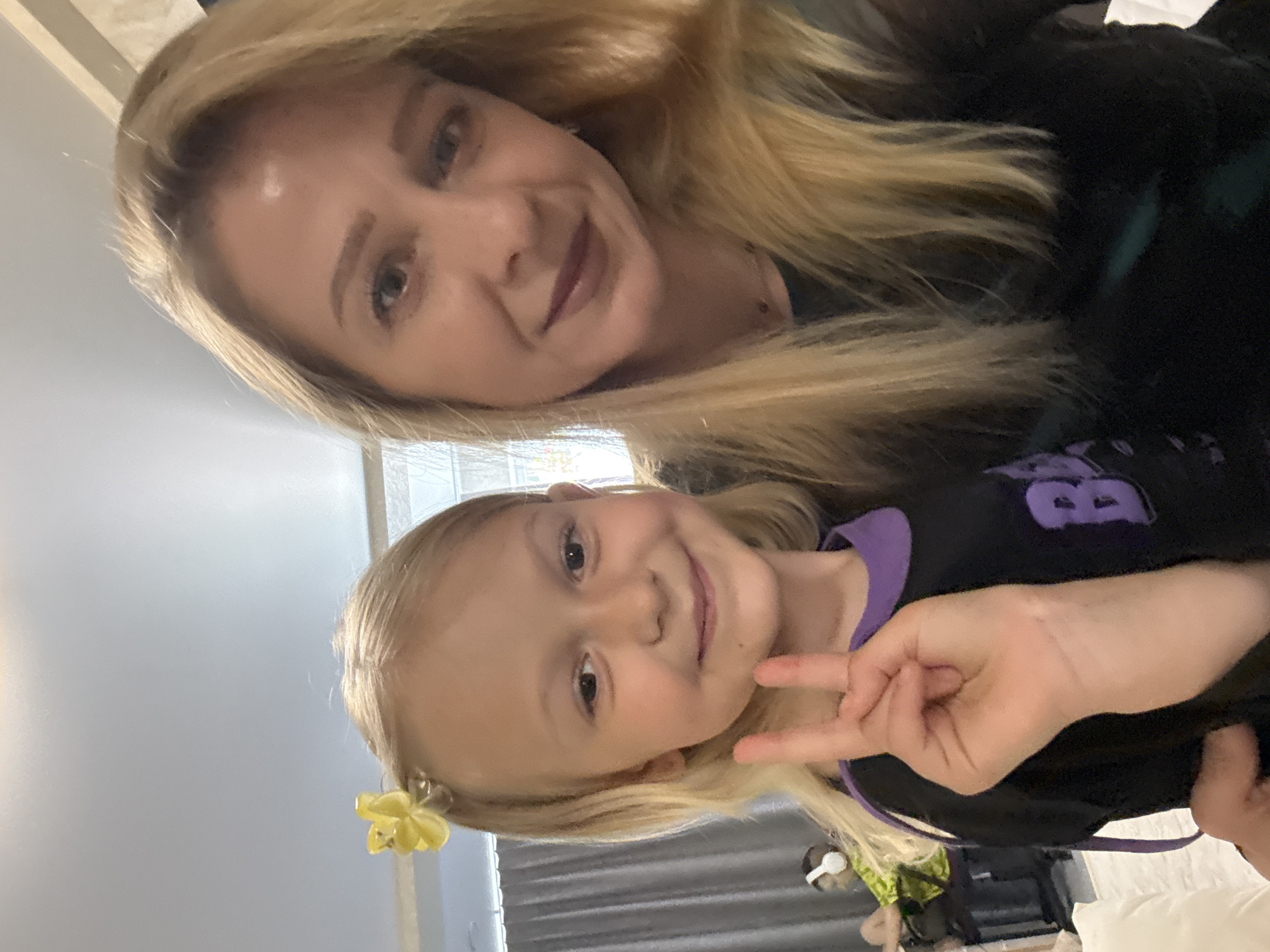 A selfie shows a woman with blonde hair posing with a young girl, also blonde, who is making a peace sign. The woman is wearing a dark top, and the girl is wearing a black and purple shirt. A yellow flower clip is in the girl's hair. The location appears to be indoors, possibly a bedroom, with a bed and other objects visible in the background. The city and state are Unknown, Unknown.
