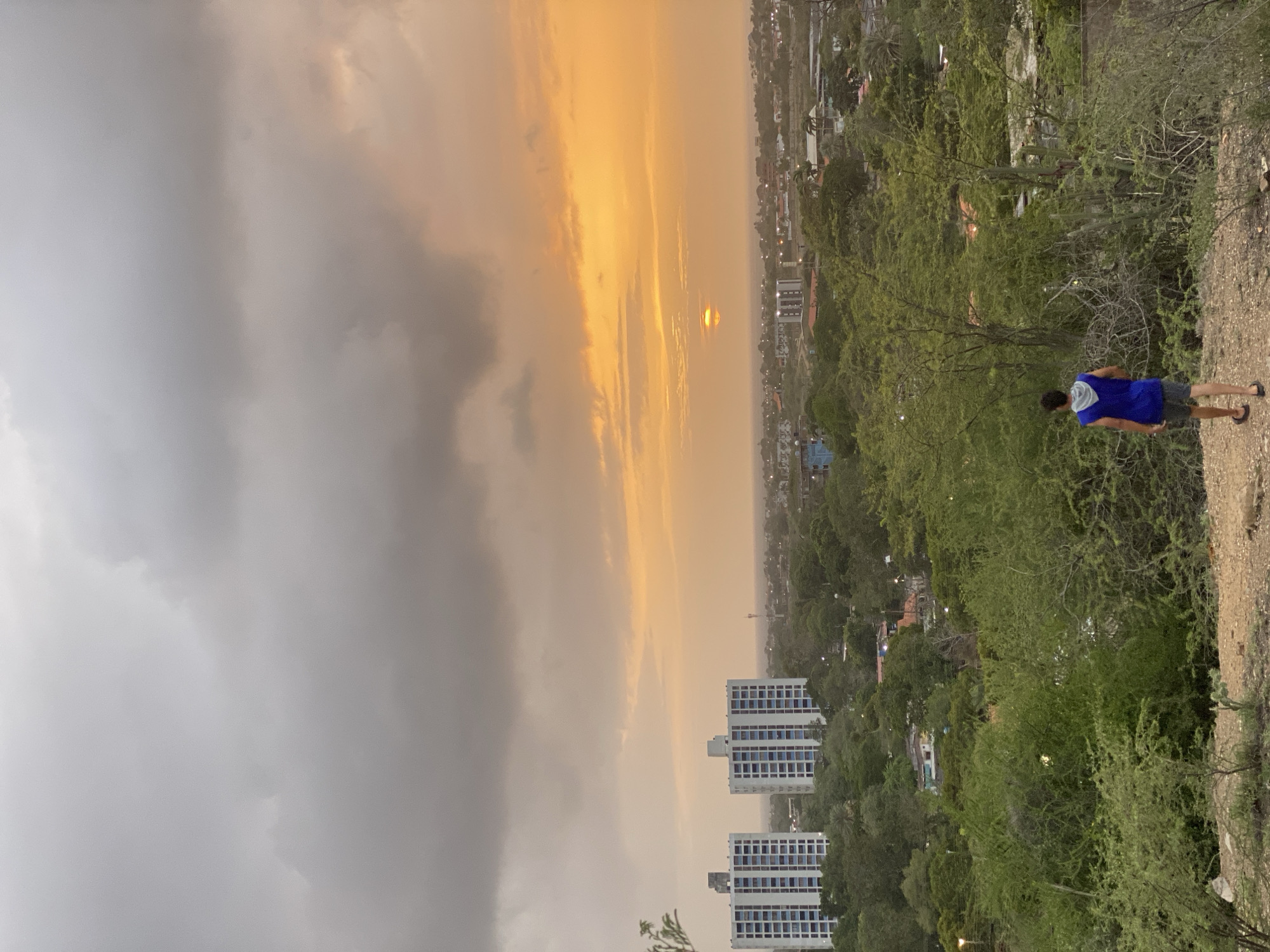 A person walks away from the camera down a dirt path on a hillside overlooking a city during sunset. The person, a young male, is wearing a blue sleeveless hoodie and dark shorts. In the background, two identical white apartment buildings stand prominently. The sky is filled with dramatic clouds, with the sun setting in a brilliant orange and yellow glow. Lush green trees and foliage cover the hillside and extend into the city. The overall scene suggests a serene evening in a coastal or tropical urban environment. This is likely Juan Griego, Venezuela, a city known for its sunsets and coastal views.