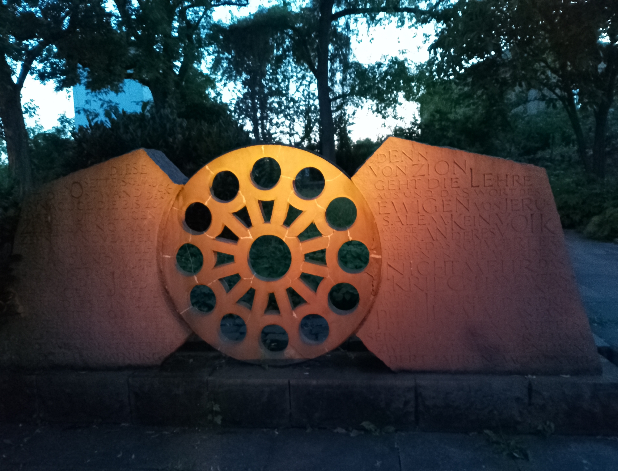 A circular, orange-toned stone sculpture with cut-out circles is centrally positioned between two larger, irregularly shaped stone slabs inscribed with text. The sculpture is illuminated from the front with warm light, casting shadows and highlighting the engraved characters on the slabs. The overall setting appears to be outdoors, with dark trees and foliage visible in the background under a twilight sky. The ground in front of the sculpture is paved with dark stone. The text on the left slab begins with "DIESE" and continues with a list of dates, while the text on the right slab includes phrases like "DENN VON ZION GEHT DIE LEHRE AUS". The condition of the sculpture and slabs appears to be weathered but intact.