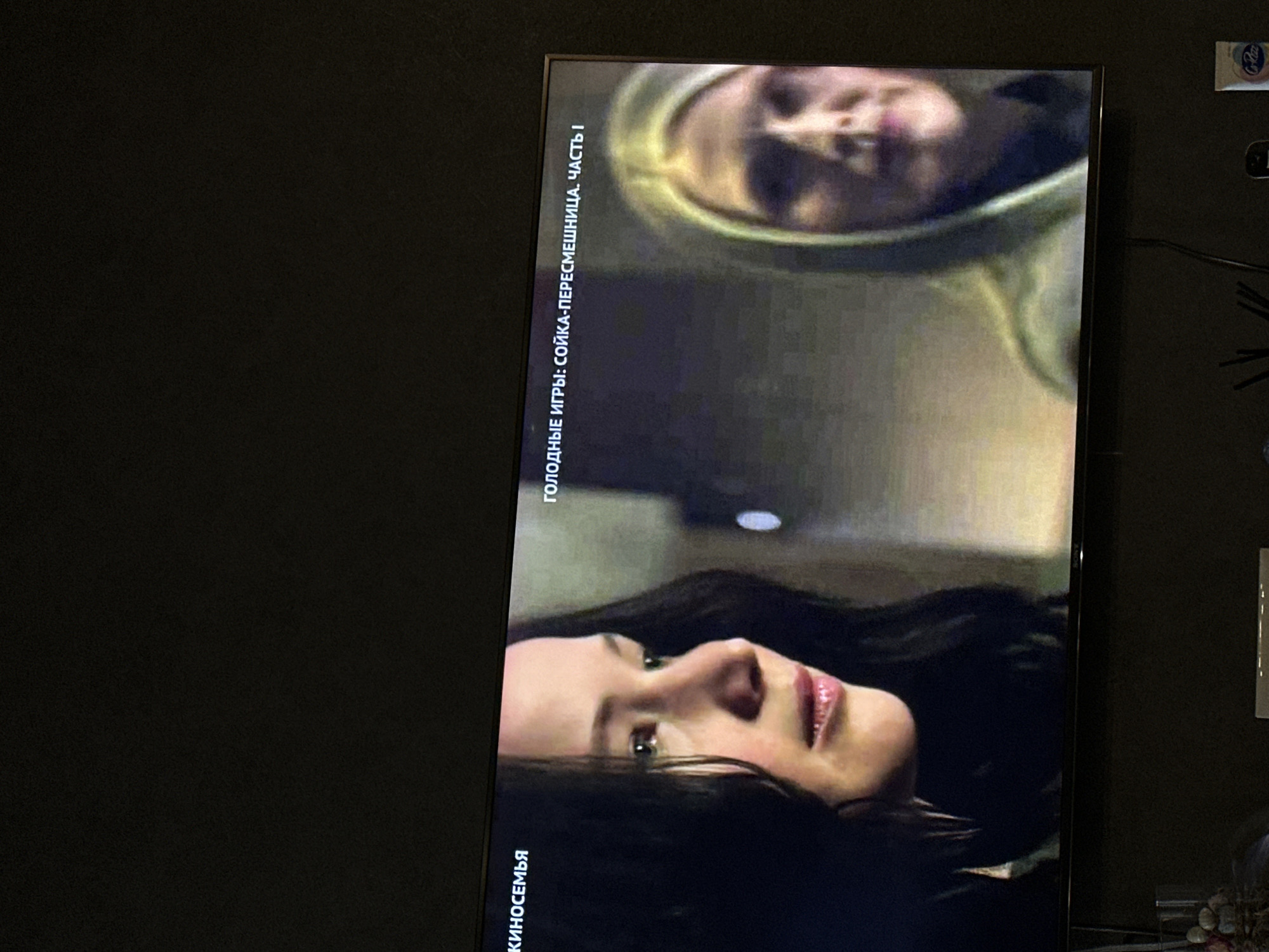 The image shows a television screen displaying a scene from a movie titled "The Hunger Games: Mockingjay Part 1". Two people are visible on the screen. Text in Russian, "ГОЛОДНЫЕ ИГРЫ: СОЙКА-ПЕРЕСМЕШНИЦА. ЧАСТЬ I" appears at the top of the screen. The television is mounted on a dark-colored wall. The wall appears to be indoors. The image does not provide information about the location or the city of Yerevan, Armenia.
