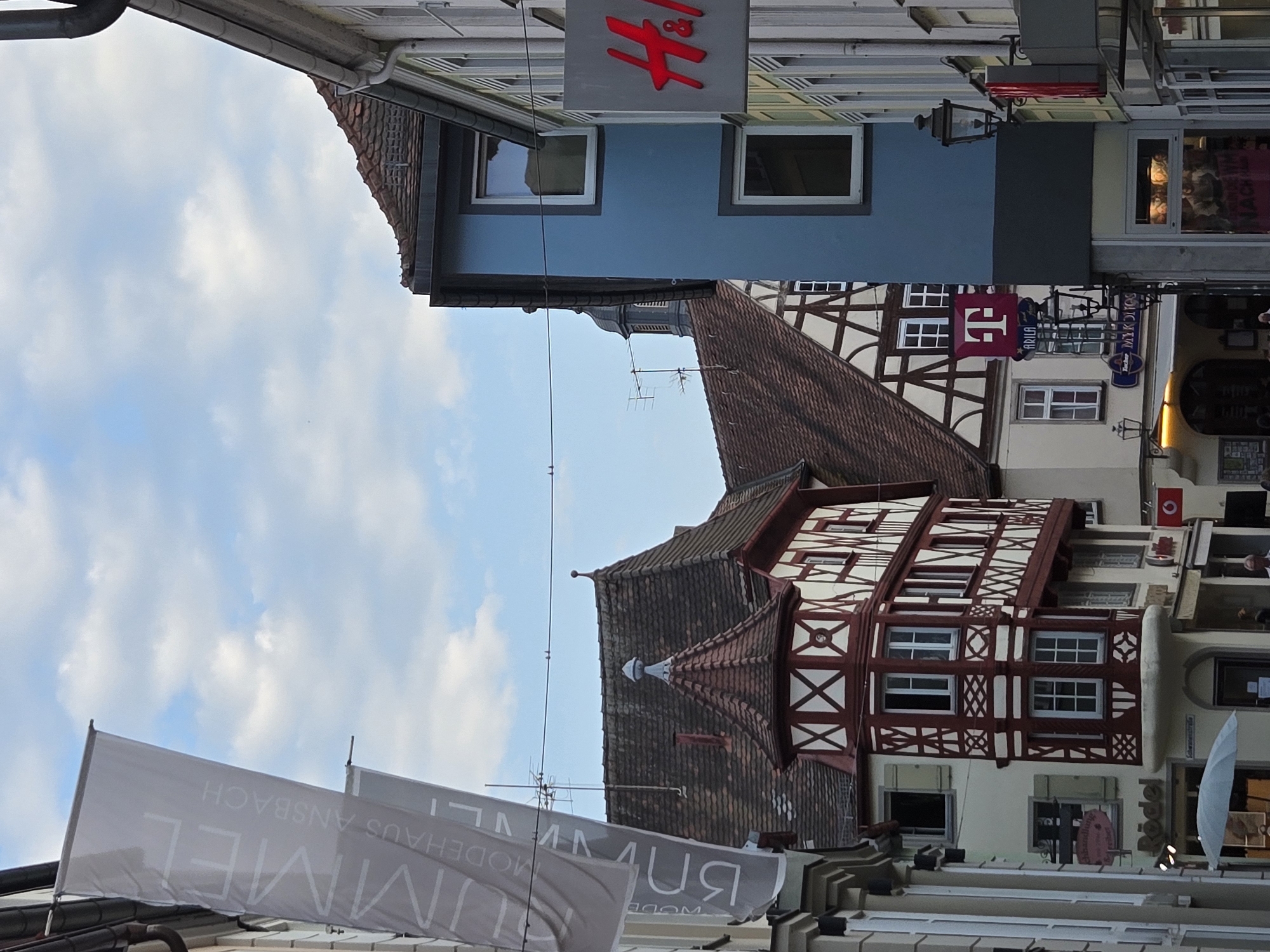 This image depicts a street scene in Ansbach, Germany, likely in the historic city center. In the foreground and left, a tall, light gray banner with dark lettering hangs vertically, displaying the words "HIMMEL MODEHAUS ANSBACH." To the right, several buildings are visible. One building features traditional half-timbered architecture with white plaster and dark wood beams, topped with a red-tiled roof. Adjacent to this, a blue building with white-framed windows stands out. Further to the right, a building with a light-colored facade has a prominent red "H&M" logo sign. A street lamp is mounted on the side of the blue building. The sky above is blue with scattered white clouds. The scene appears to be from a shopping district, with commercial signage visible on some of the buildings.
