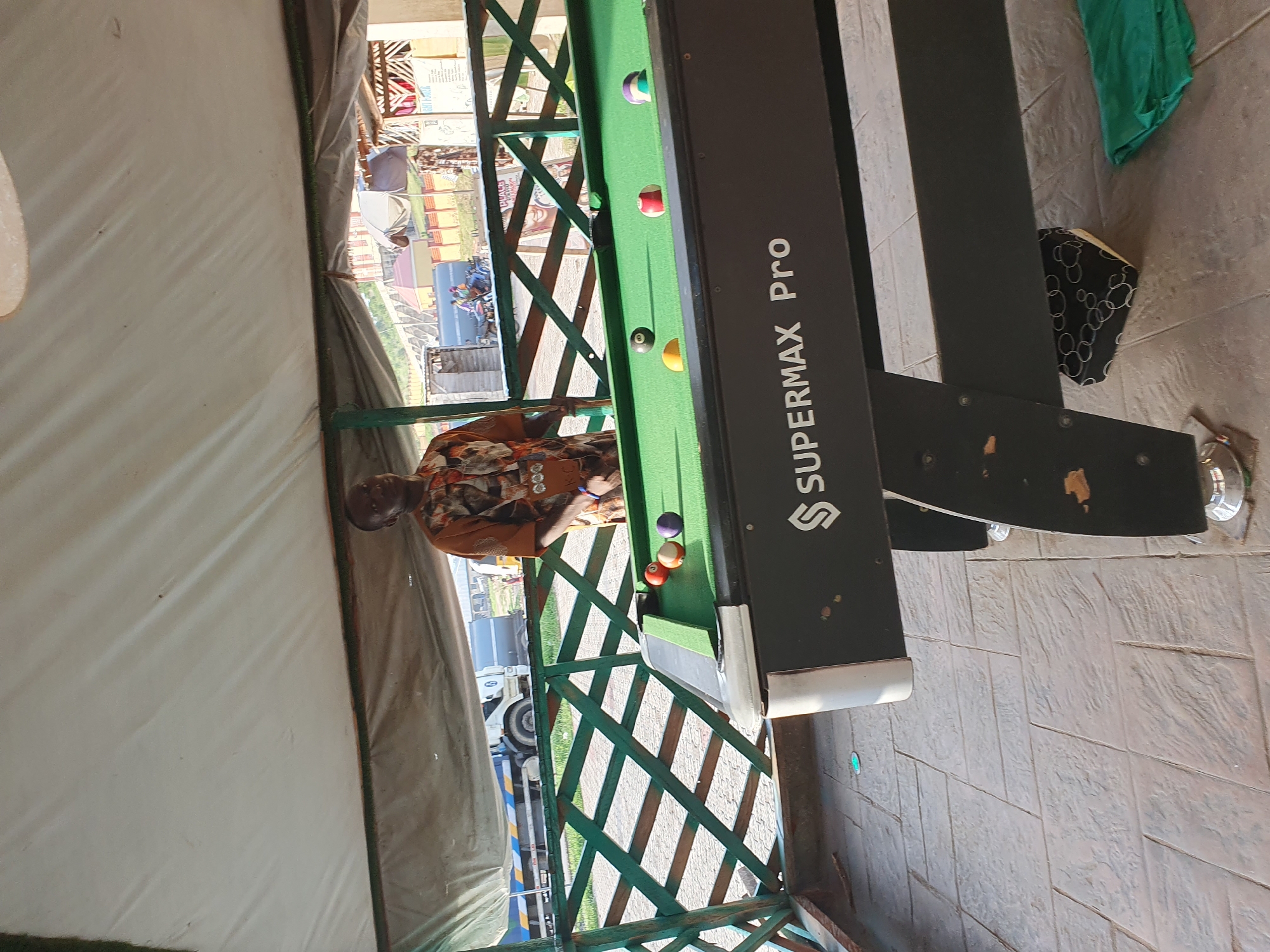 A man stands behind a pool table under a white awning in Lokoja, Nigeria. The pool table is green with several balls on it, including a red and white striped ball, a purple ball, an orange ball, a yellow ball, and a black ball. The man is wearing a patterned shirt. Behind the pool table is a green latticed fence. Beyond the fence, a truck and some buildings are visible. The ground is paved with large rectangular stones.