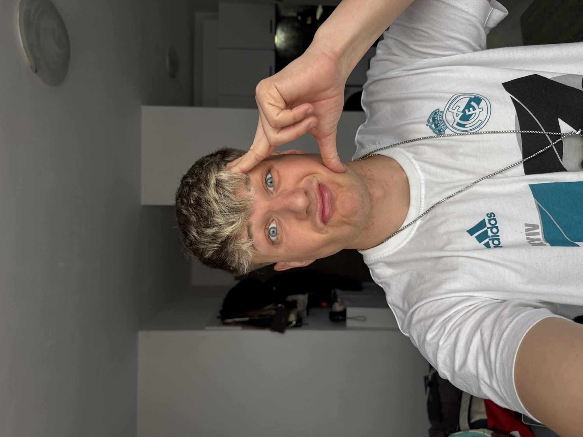 A young man poses indoors wearing a white t-shirt with the Real Madrid club emblem and Adidas logo. He is making a gesture with his hand near his face. The location is not discernible from the image, making it impossible to determine if the scene is in Plovdiv, Bulgaria.