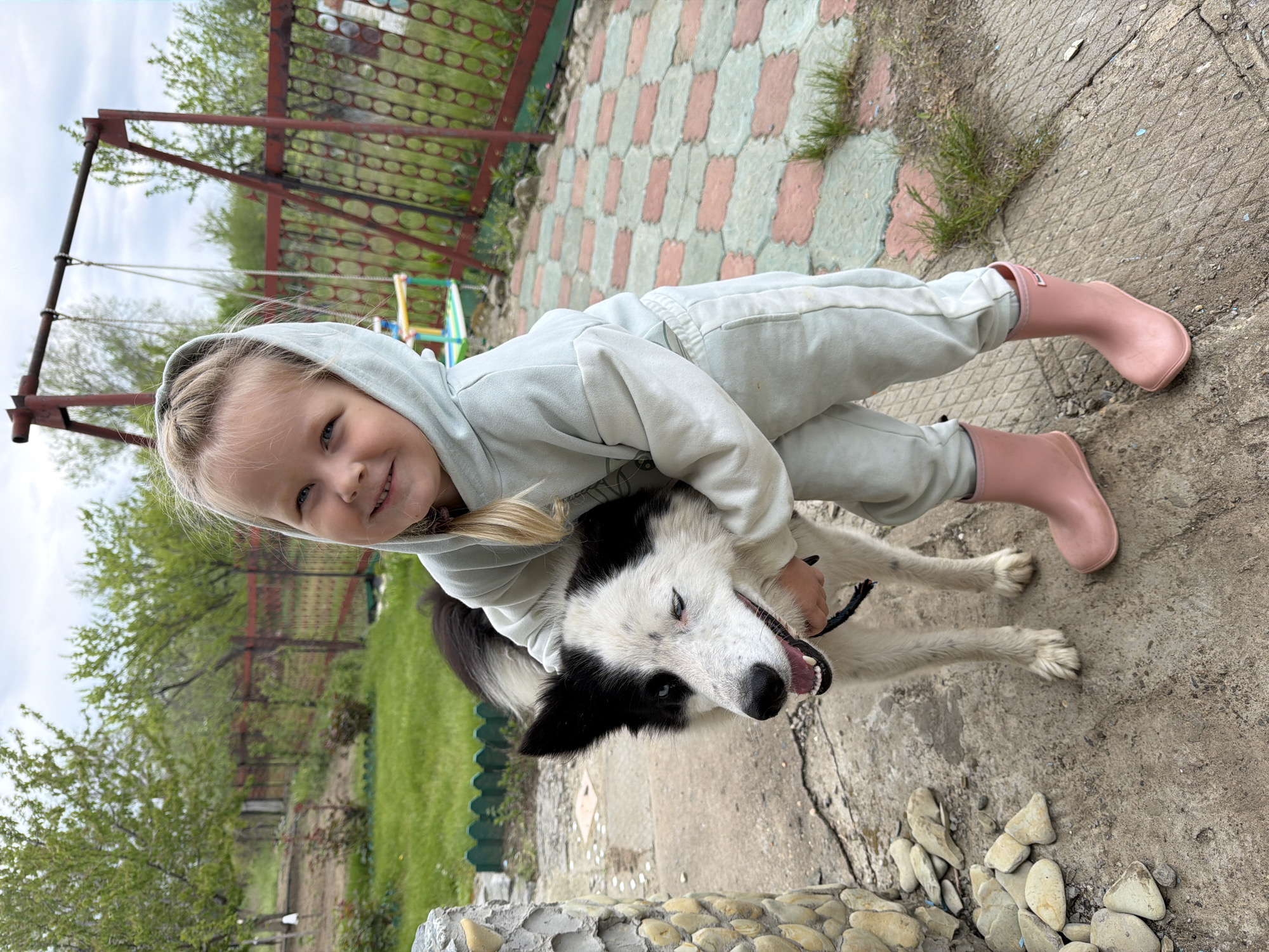 A young girl in a light-colored hooded jumpsuit and pink boots is hugging a white and black dog outside. The dog appears to be smiling. The background includes a swing set, a fence, patterned paving stones, and a concrete area with stones at the edge. The location is Kamyshin, Russia.
