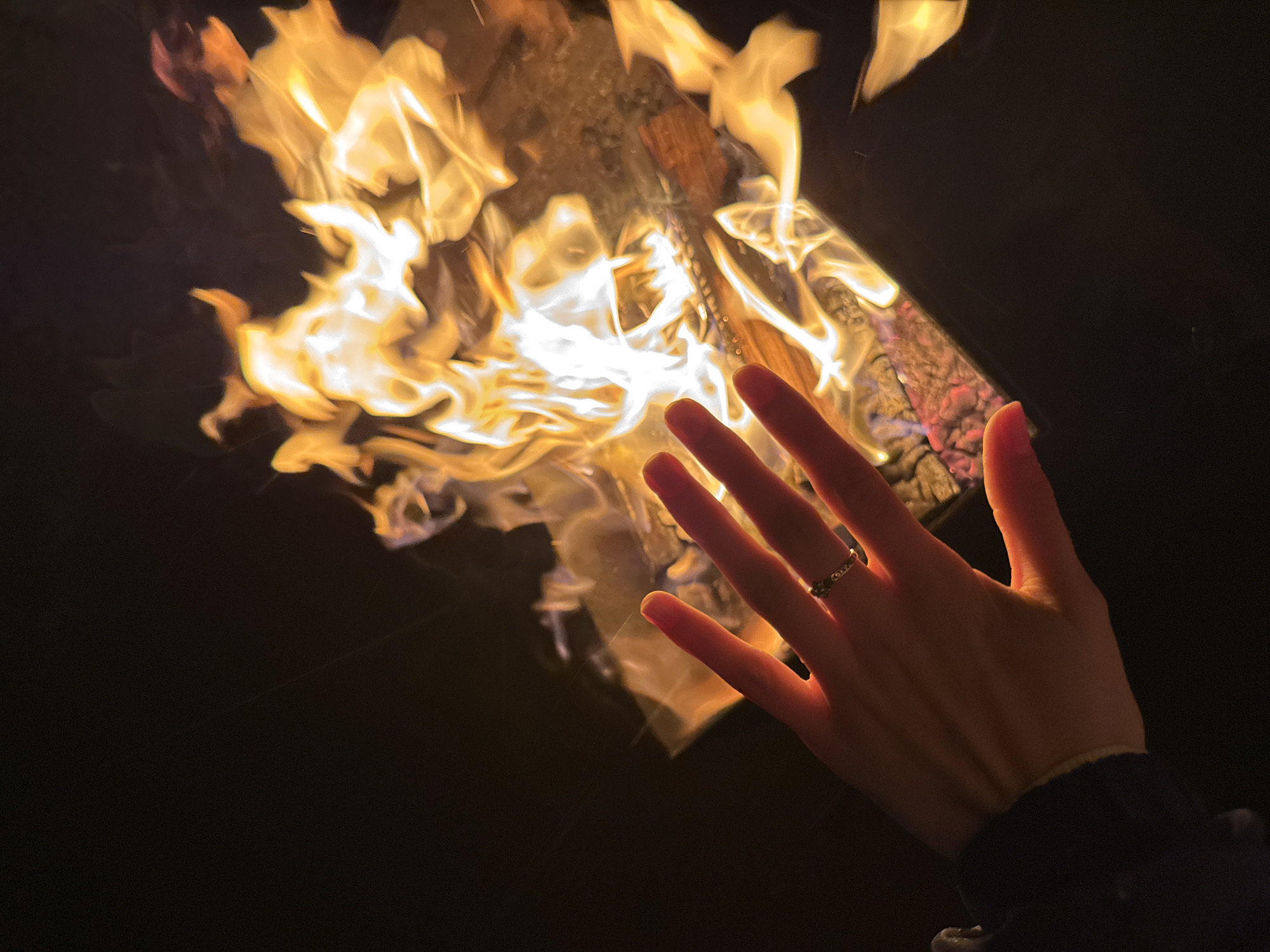 A close-up shot shows a person's hand near a bonfire. The hand is pale and illuminated by the firelight, with a ring visible on one finger. The bonfire consists of burning wood, with flames reaching upwards. The surrounding environment is dark, suggesting the scene is at night. The location of Mayak, Russia is not evident in the image.