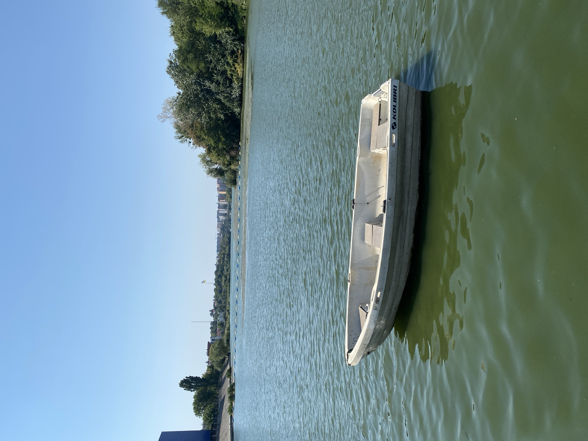 The image shows a small, white, rowboat named "Kolibri" floating on a green body of water, likely a river or lake. The boat is empty and stationary. The water surface is rippled. In the background, a tree-lined shore is visible, with buildings and a cityscape beyond. A Ukrainian flag is planted on a hill in the distance. The sky is clear and blue. This scene is likely located in Vinnytsia, Ukraine, possibly on the Southern Bug River.