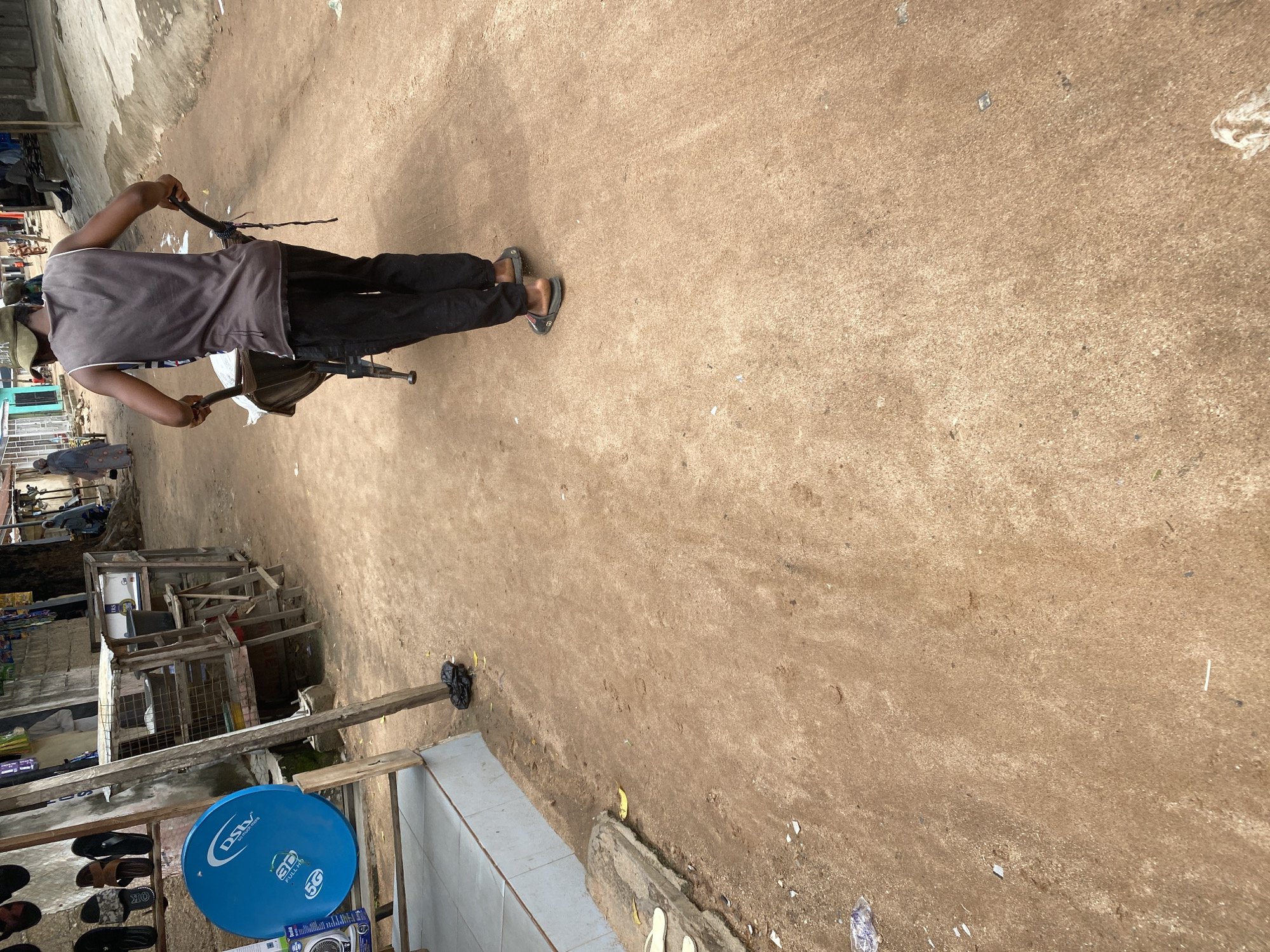 A man in a grey tank top and black pants, wearing flip-flops, is pushing a wheelbarrow on a dirt road in Jalingo, Nigeria. The wheelbarrow contains some white material. In the background, there are buildings, a tree, and other people walking. To the left, there is a blue satellite dish and a stall selling footwear. The environment appears to be an outdoor market or street scene.