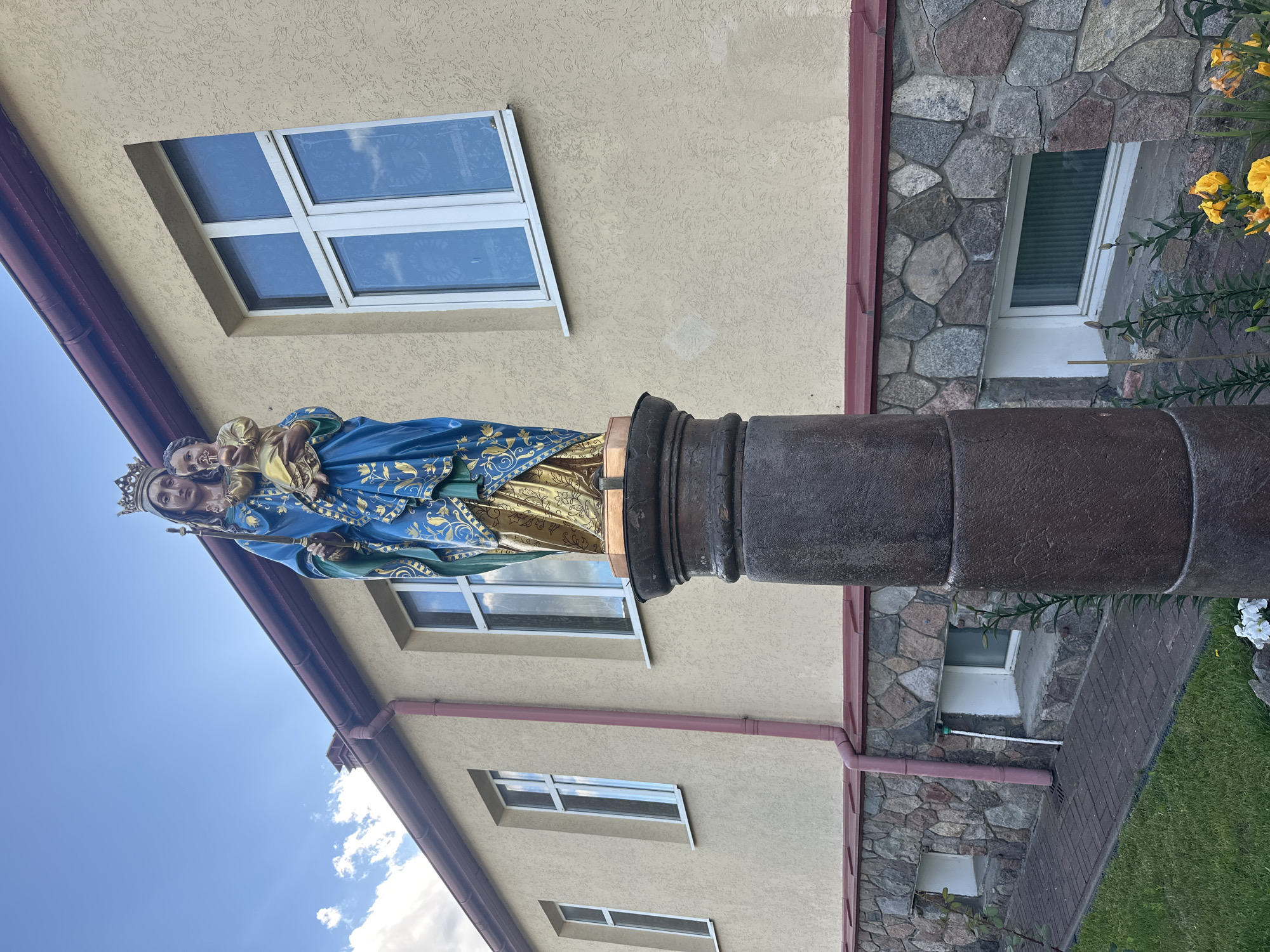A statue of the Virgin Mary holding the infant Jesus stands on a stone pedestal in front of a building with a textured, light-yellow facade. The statue is brightly colored, with Mary wearing a blue robe adorned with gold patterns and a golden crown. The infant Jesus is also depicted in gold. The building has several windows with white frames and what appear to be reflective panes, and a dark red roofline. The lower portion of the building is constructed with irregular, multi-colored stones. A red downspout runs vertically along the facade. The ground in front of the building is a paved walkway with a brick pattern, bordered by a grassy area. The sky above is blue with scattered white clouds. The image does not provide any specific information about the city of Minsk, Belarus.