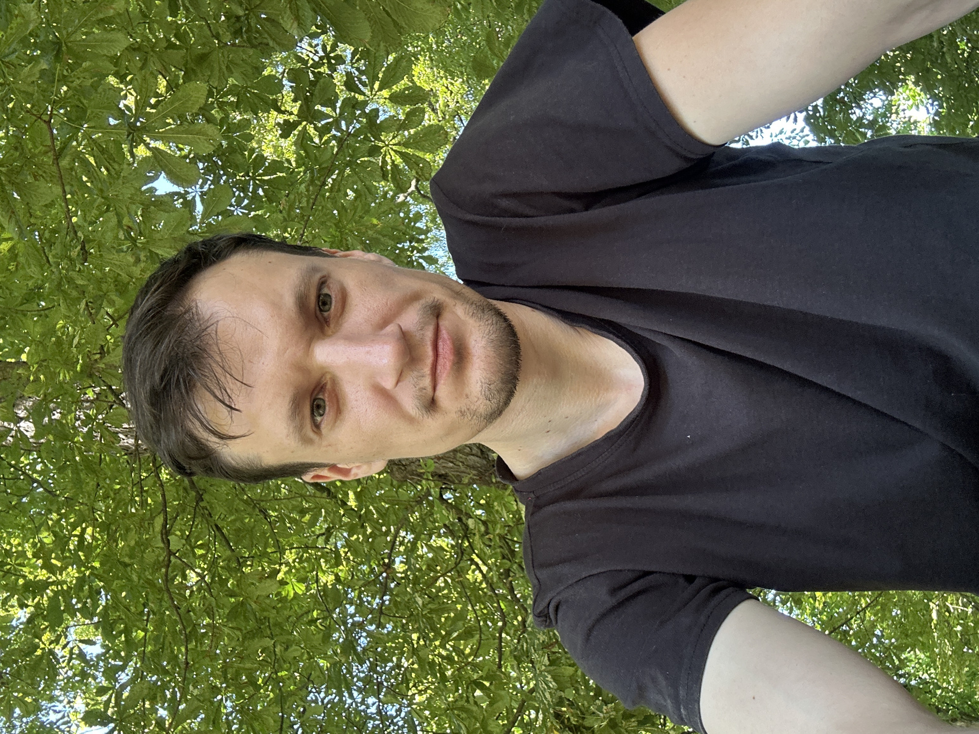 A selfie of a man with brown hair, a light beard, and green eyes is taken outdoors. He is wearing a black t-shirt and is positioned in front of a backdrop of lush green leaves and a tree trunk. The lighting suggests it is daytime. The man's gaze is directed at the camera. There is no indication of the specific location in Makiv, Ukraine.
