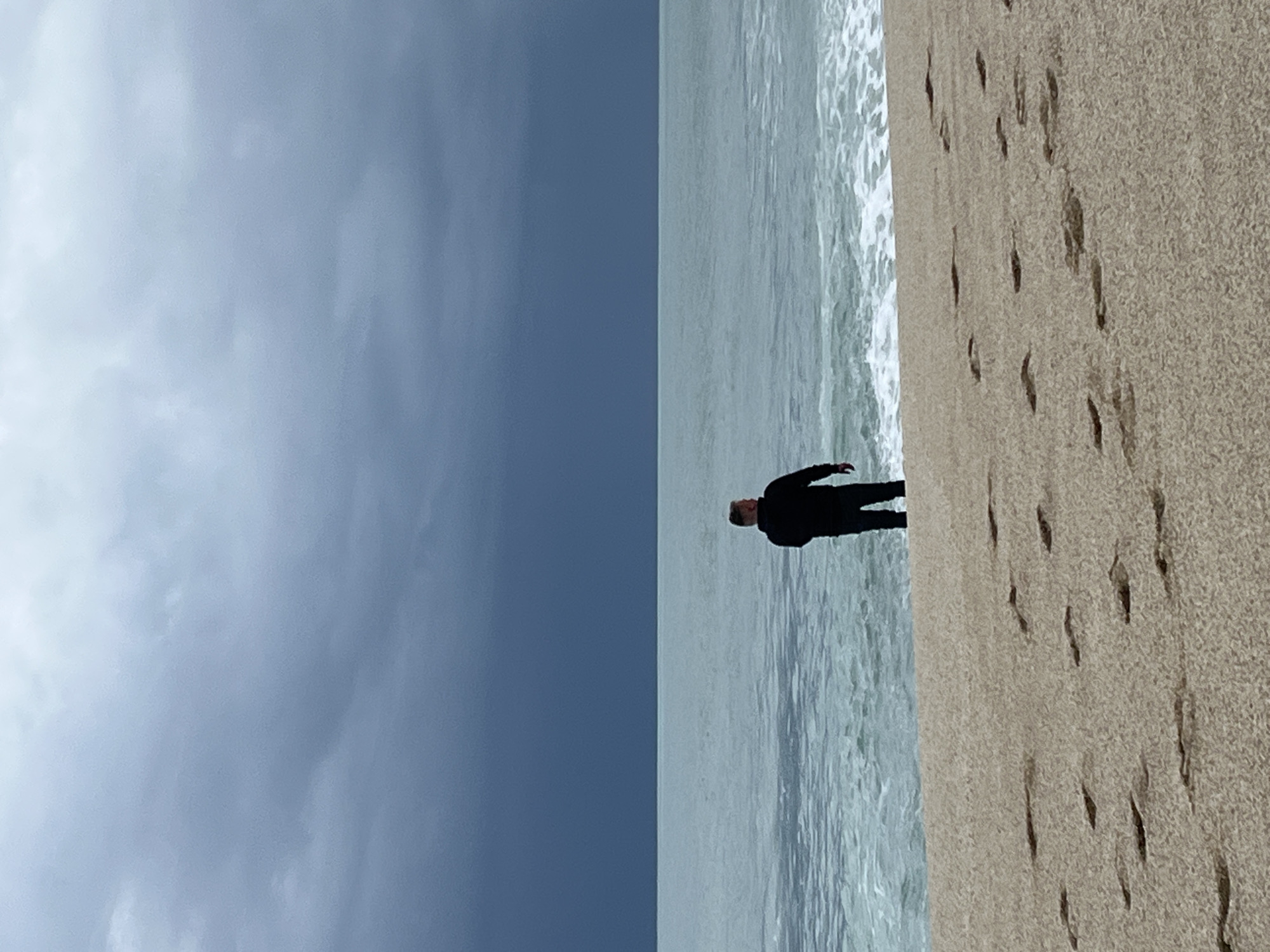 The image shows a beach scene in Alanya, Turkey. The sky is overcast. A person is standing in the water at the edge of the beach, near the line of breaking waves. There are footprints in the sand along the beach. The sand is light brown.