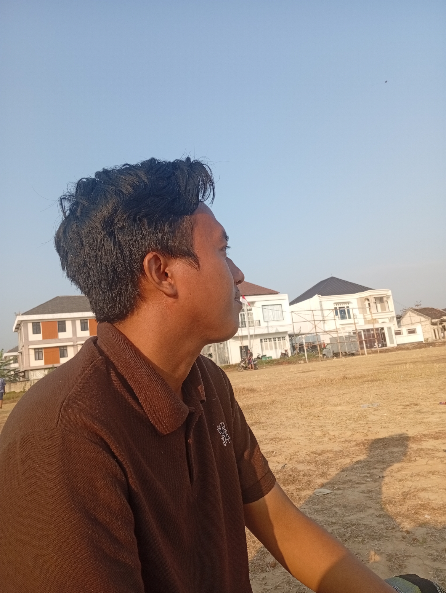 A young man with dark hair is shown in profile, looking to the right, against a bright, clear sky. He is wearing a brown polo shirt. In the background, several modern houses are visible, some of which appear to be under construction. The ground in front of the houses is a dry, yellowish field, likely an open area in the city of Sarangmeduro, Indonesia. The lighting suggests it is daytime, possibly late afternoon, given the shadows.